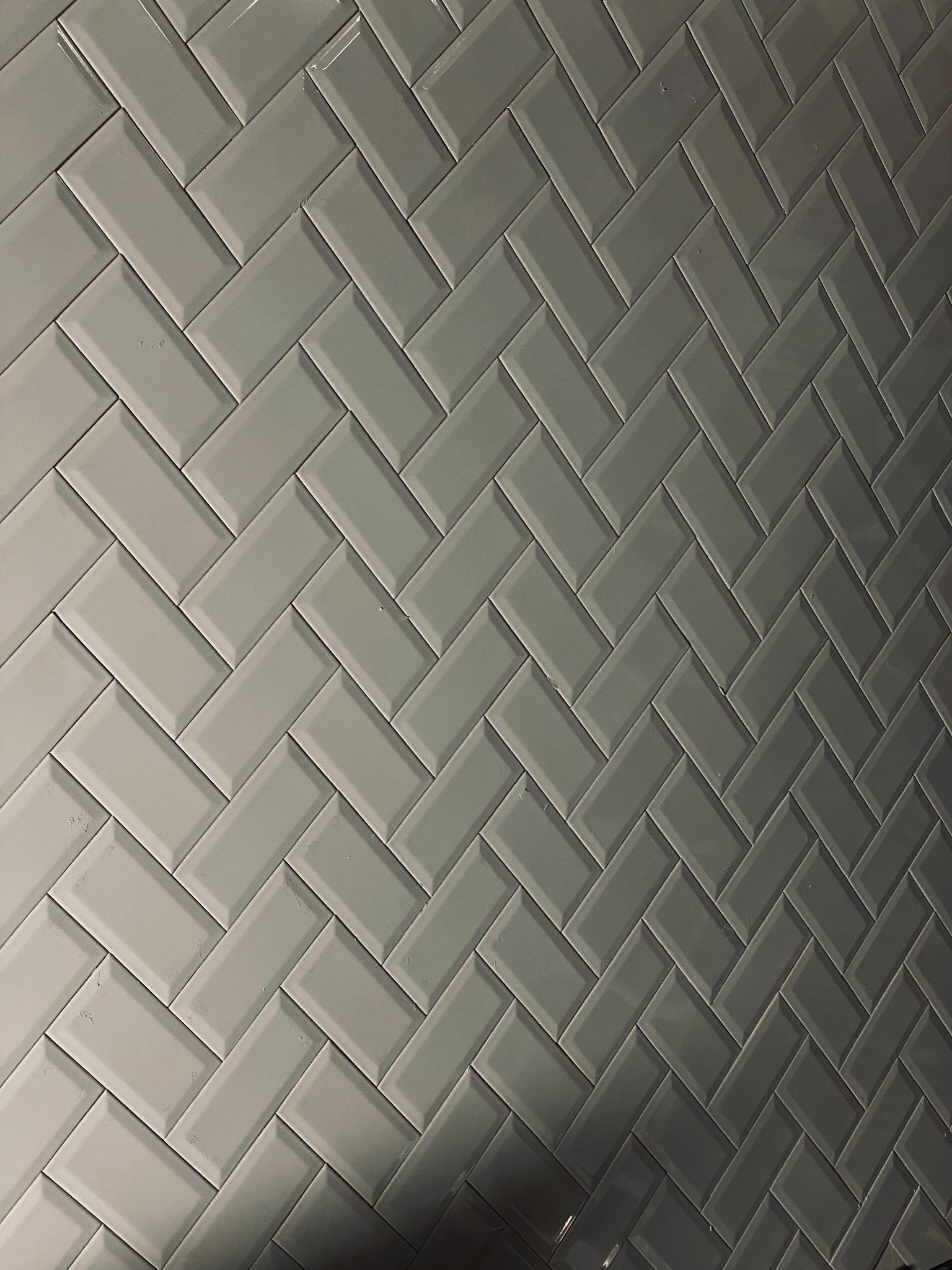 Ściana wyłożona błyszczącymi, szarymi płytkami typu metro ułożonymi w jodełkę, widoczne delikatne refleksy światła.