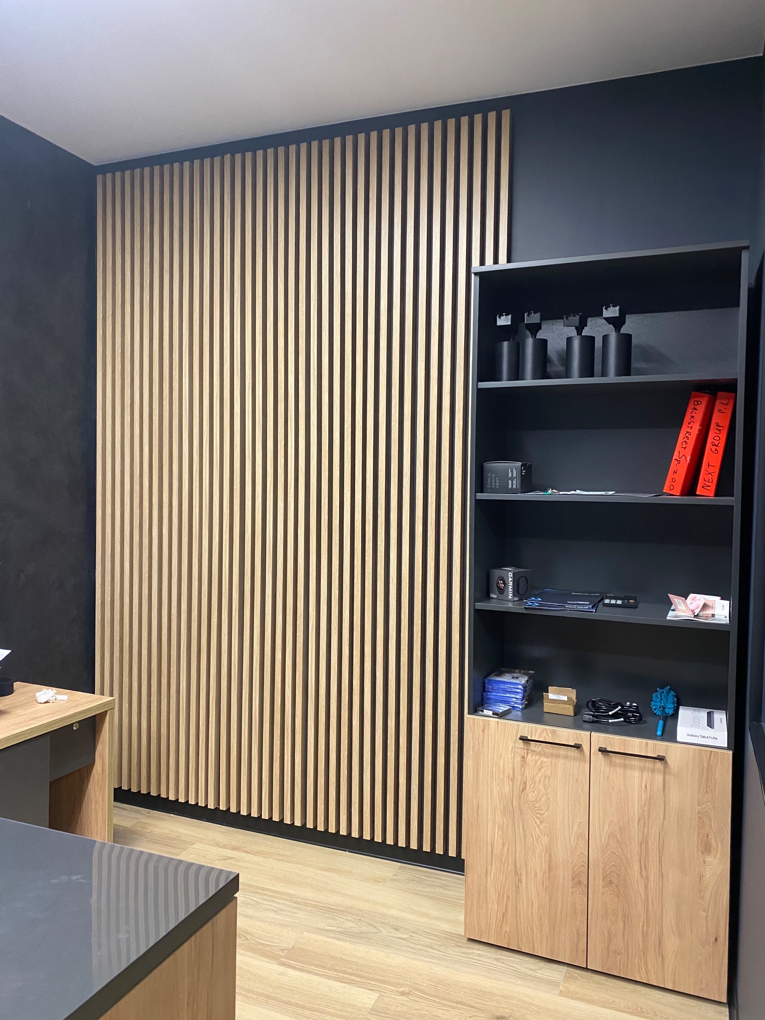 Ściana z drewnianych lameli w biurze, ciemna ściana, regał z szarymi półkami i drewnianymi drzwiczkami na dole, widoczne akcesoria biurowe i dekoracje na półkach, podłoga w jasnym odcieniu drewna.