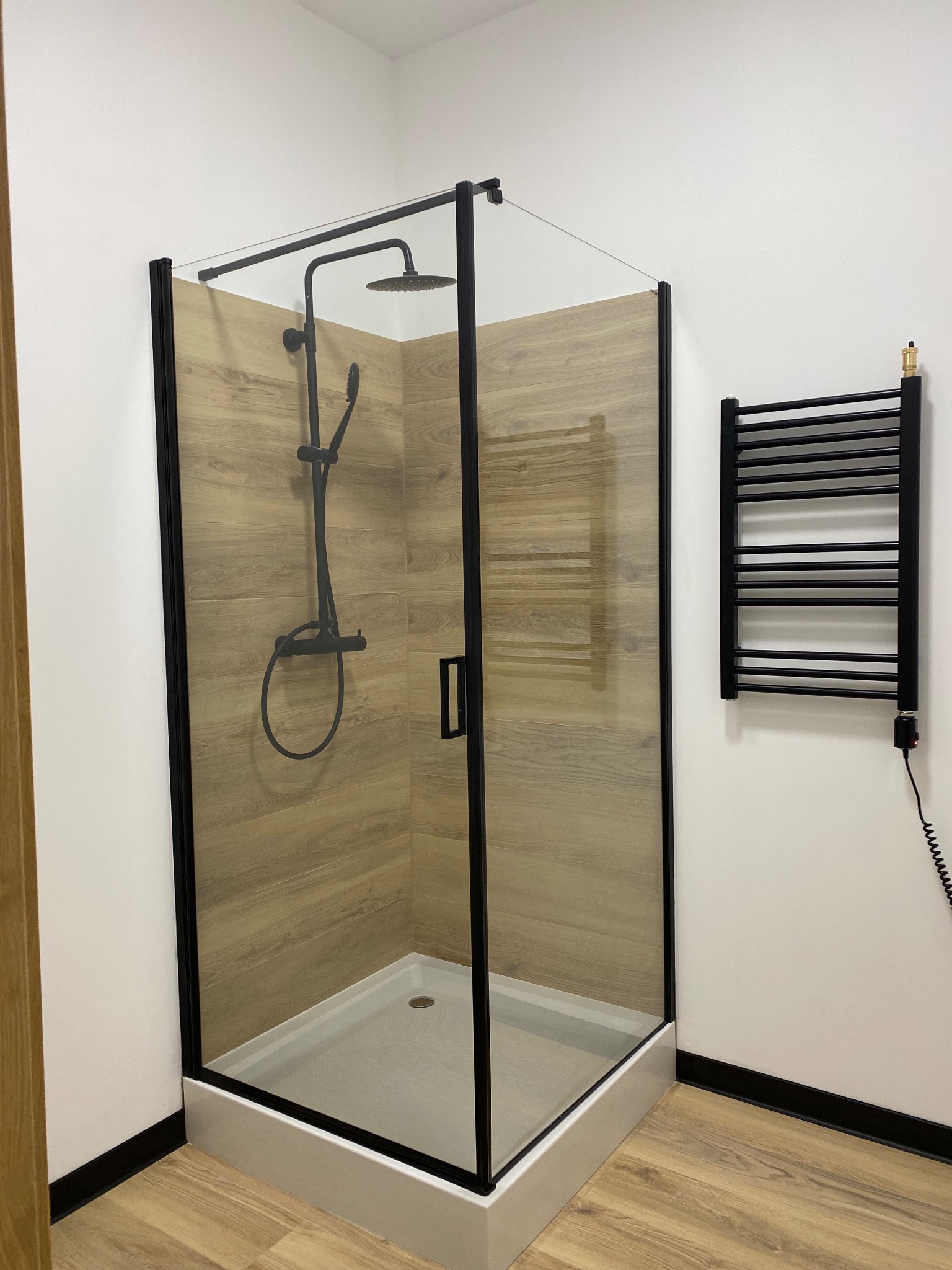 Nowoczesna kabina prysznicowa z czarną armaturą i grzejnikiem drabinkowym, wykończona płytkami imitującymi drewno, widok z boku.