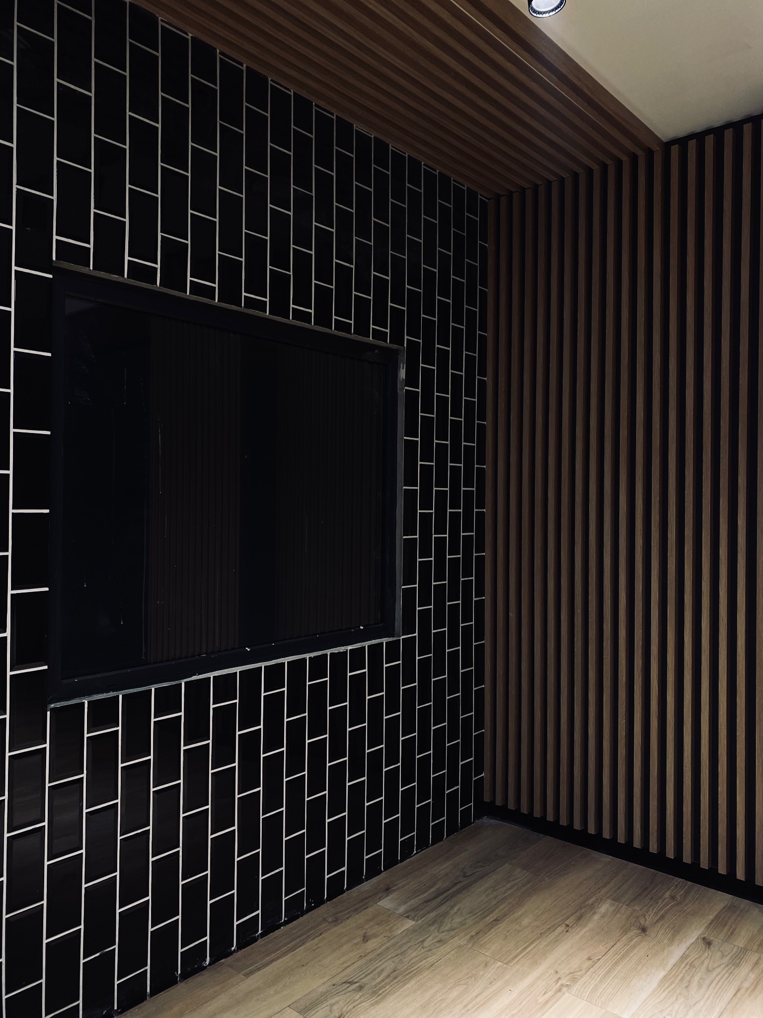 Nowoczesna łazienka z czarnymi, błyszczącymi płytkami ułożonymi w pionowy wzór cegły, kontrastującymi białymi fugami i drewnianymi panelami ściennymi, podłoga z jasnego drewna.