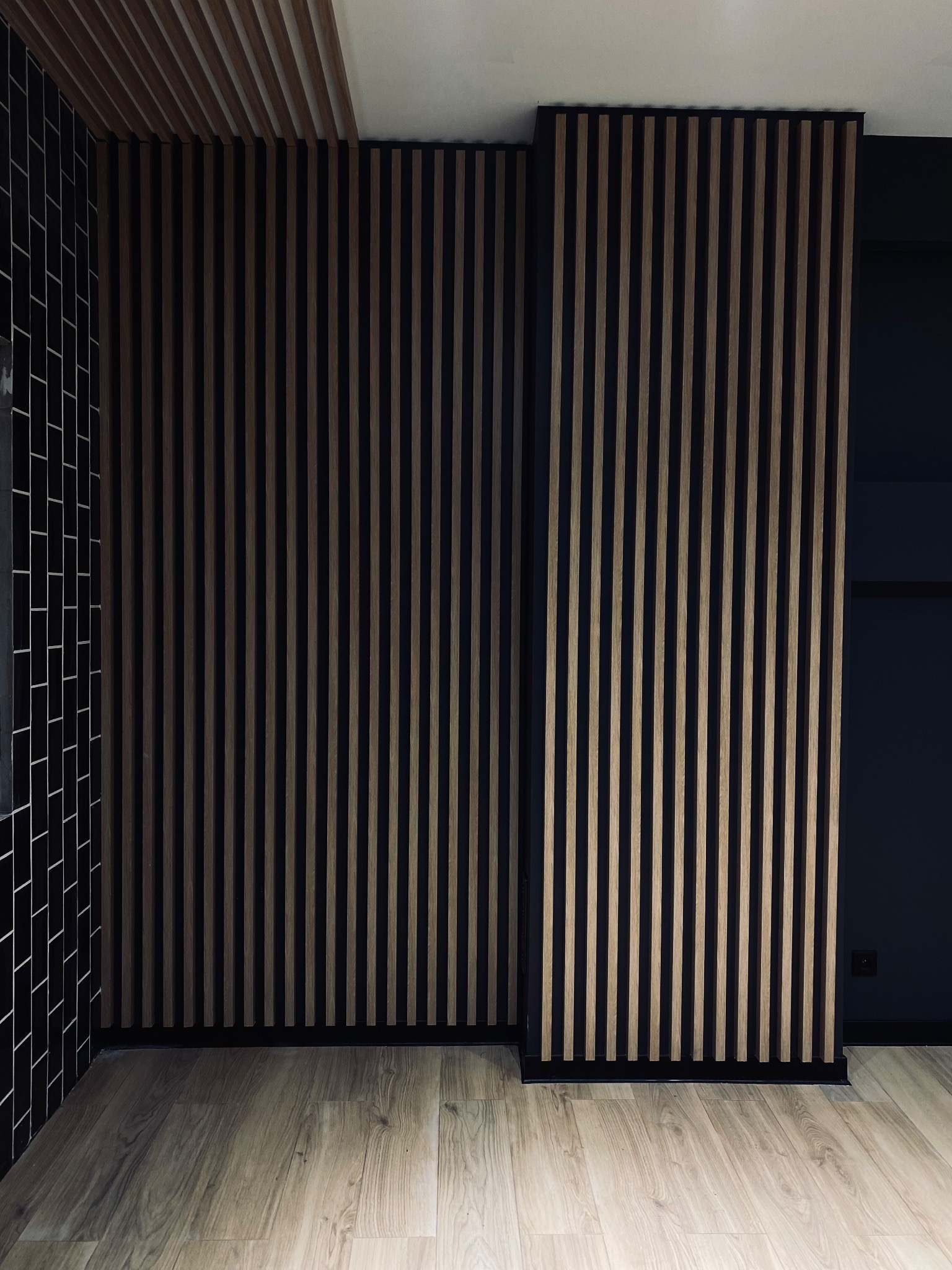 Nowoczesne wnętrze z drewnianymi lamelami na ścianie i suficie, czarne płytki na ścianie po lewej, jasna drewniana podłoga, minimalistyczny design.