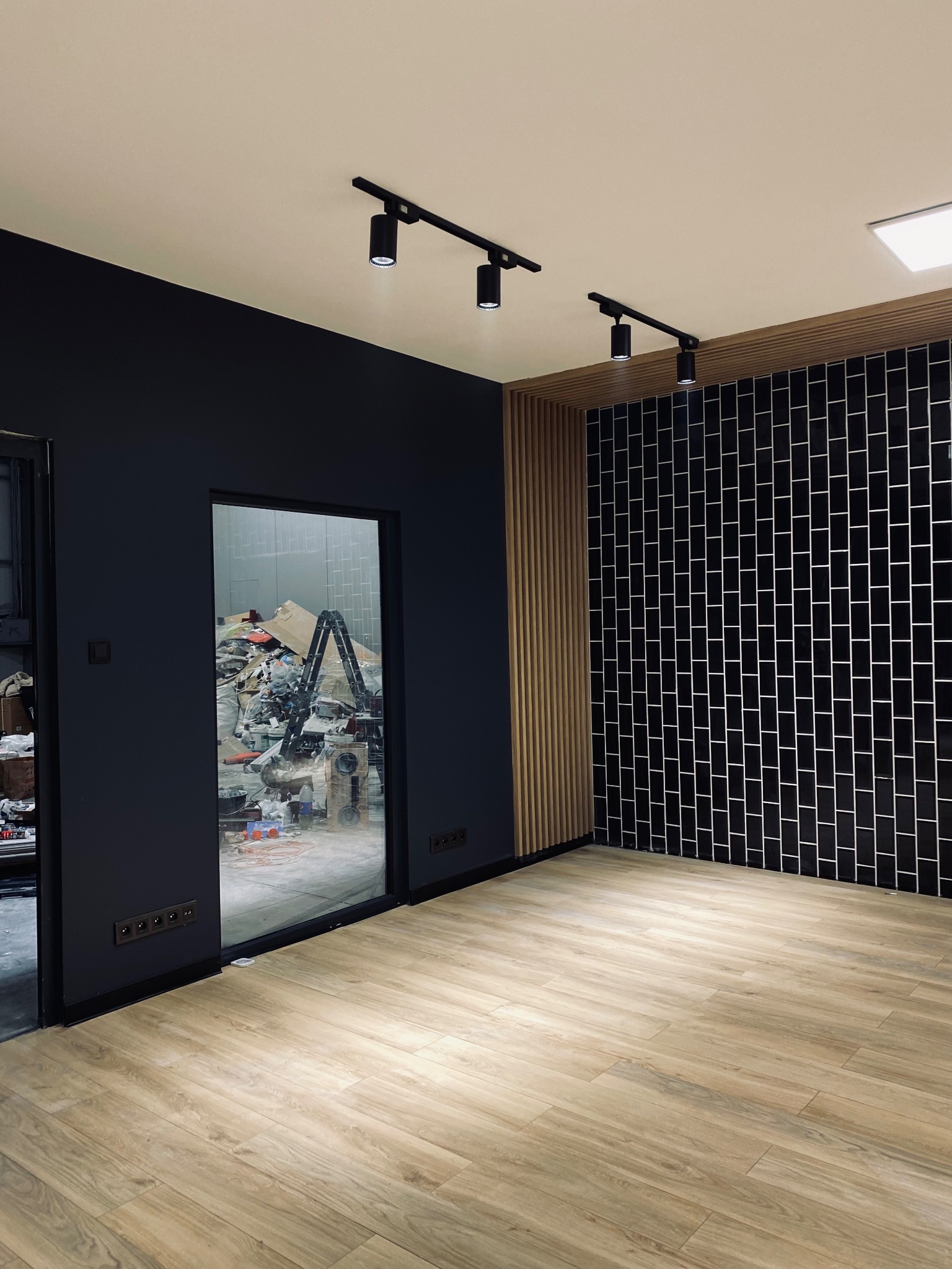 Wnętrze pokoju z parkietem, czarną ścianą i ścianą wyłożoną czarnymi, podłużnymi płytkami; w tle widoczny bałagan po pracach remontowych i drabina.