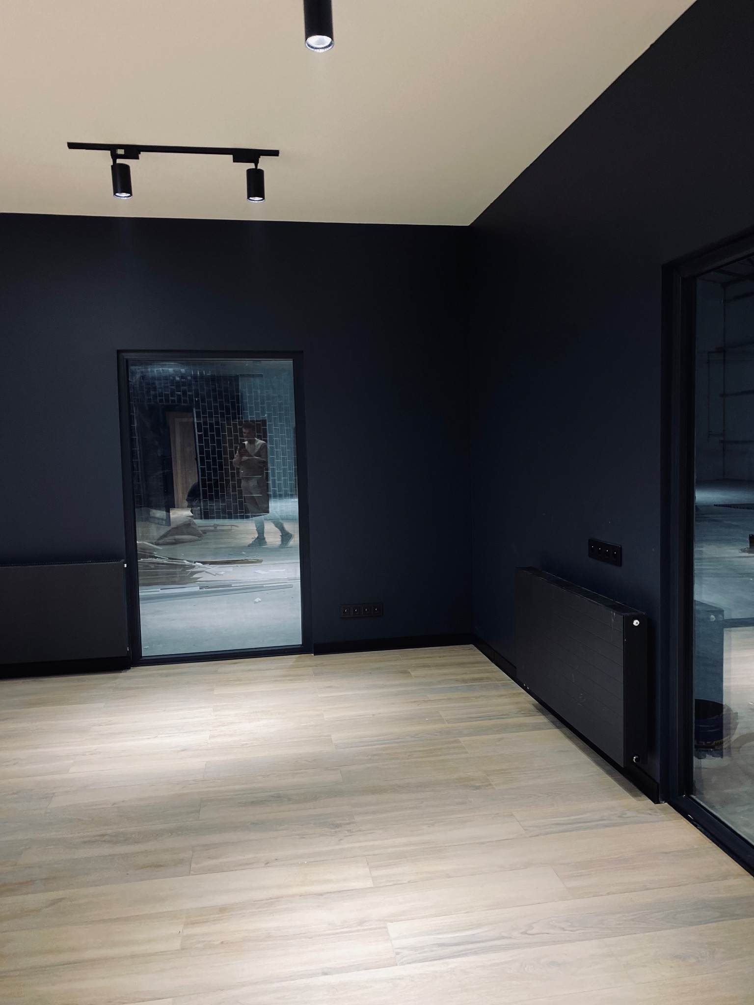 Nowoczesne wnętrze biurowe z ciemnymi ścianami, jasną podłogą imitującą drewno, czarnymi grzejnikami i oświetleniem punktowym na szynoprzewodzie.