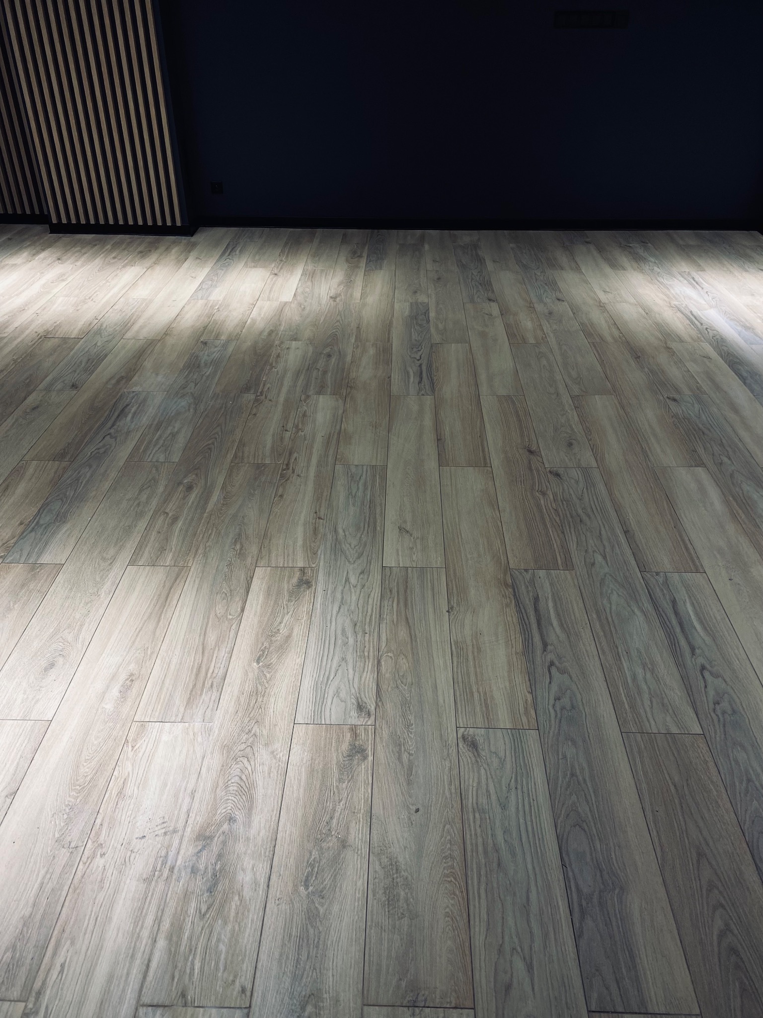 Ułożone panele podłogowe w odcieniach szarości, z widocznym fragmentem ściany w ciemnym kolorze i elementami dekoracyjnymi na suficie.