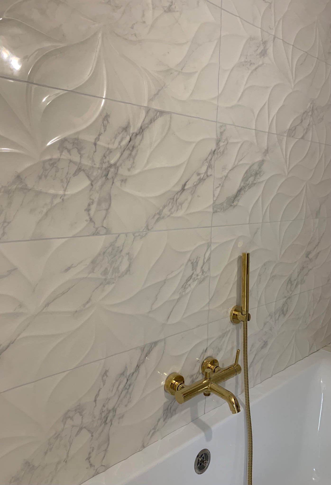 Białe, trójwymiarowe płytki imitujące marmur na ścianie łazienki, z zamontowaną złotą baterią i słuchawką prysznicową.