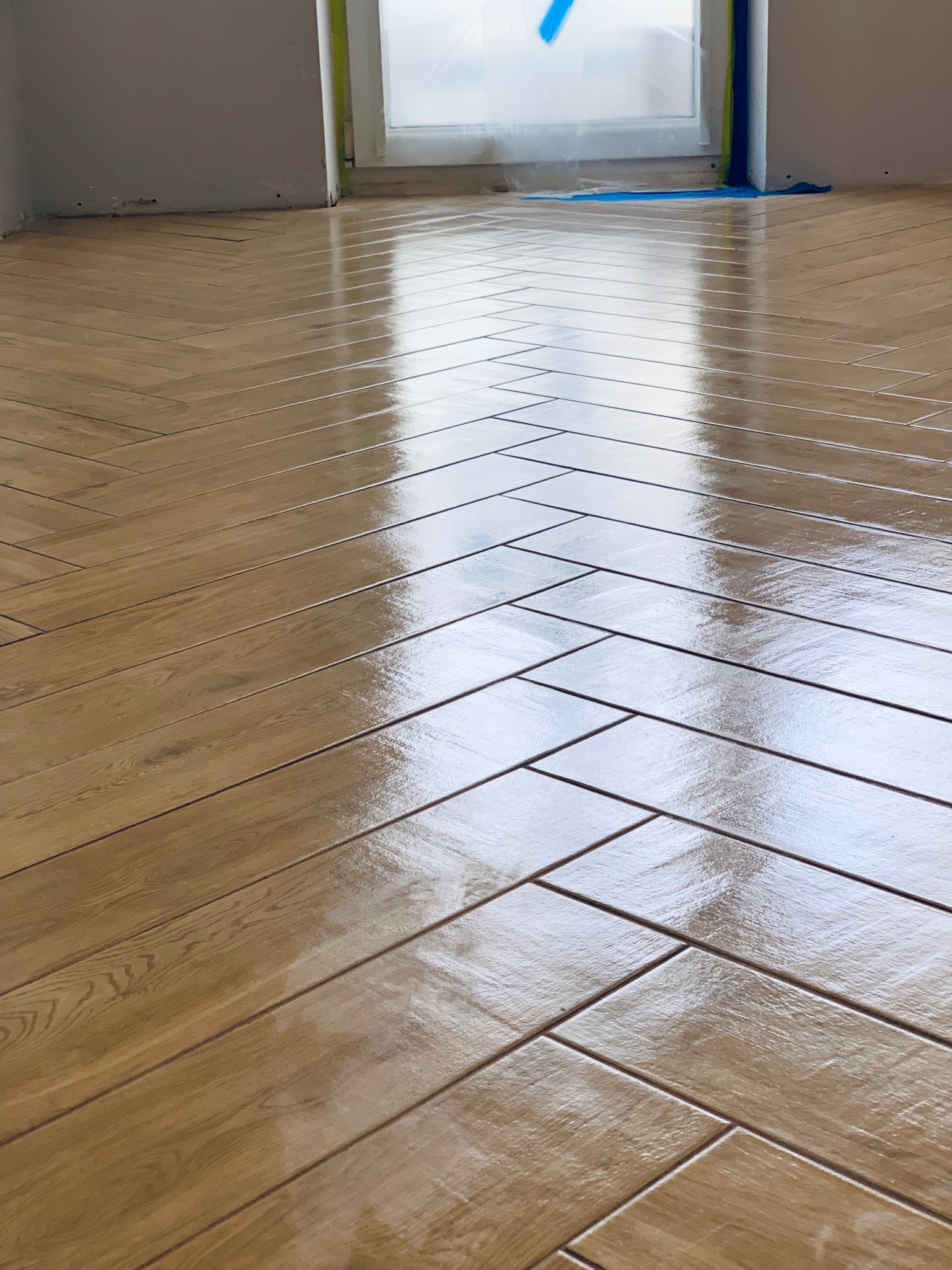 Ułożona, błyszcząca, drewnopodobna podłoga z paneli w jodełkę, zabezpieczona folią okienna rama.