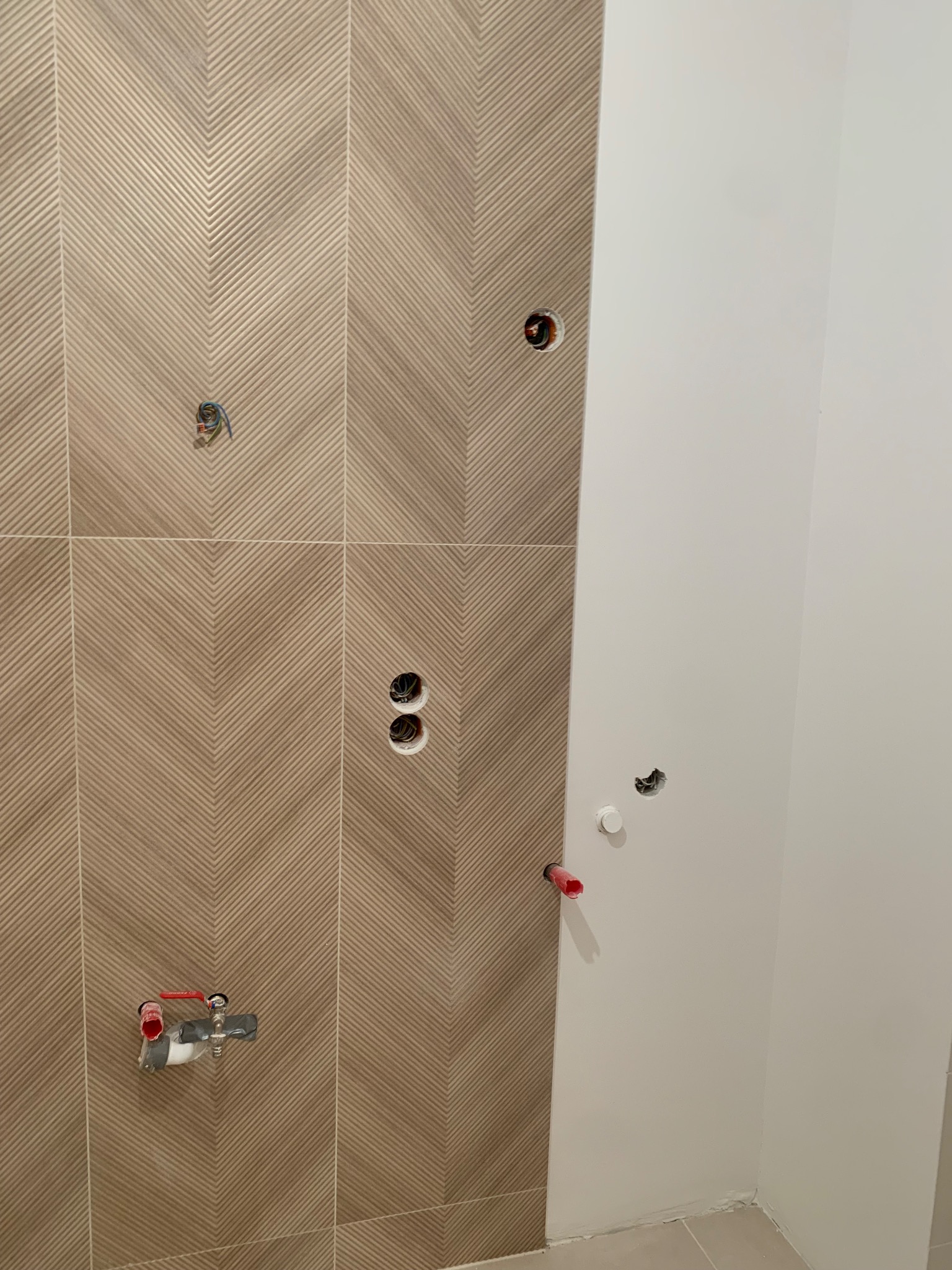 Ściana łazienki w trakcie prac wykończeniowych: ułożone płytki z wzorem chevron, widoczne otwory na instalacje wodne i elektryczne, fragment pomalowanej ściany.