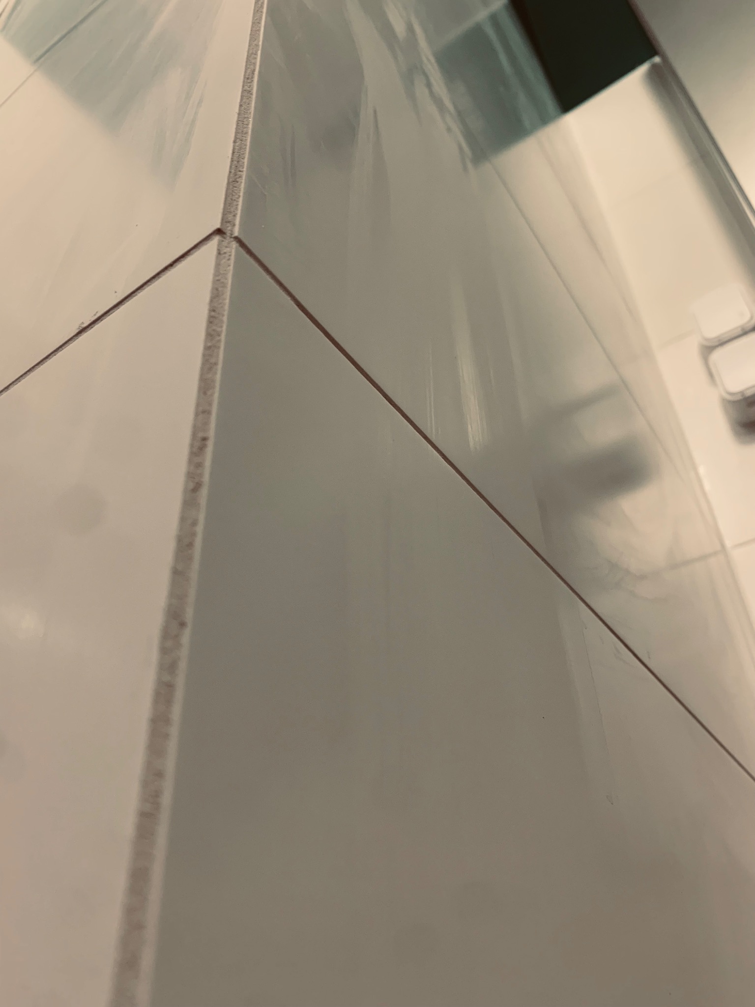 Błyszczące, białe płytki ceramiczne ułożone pod kątem, z widocznymi fugami i odbiciem światła, sugerujące świeżo wykończoną powierzchnię w łazience.