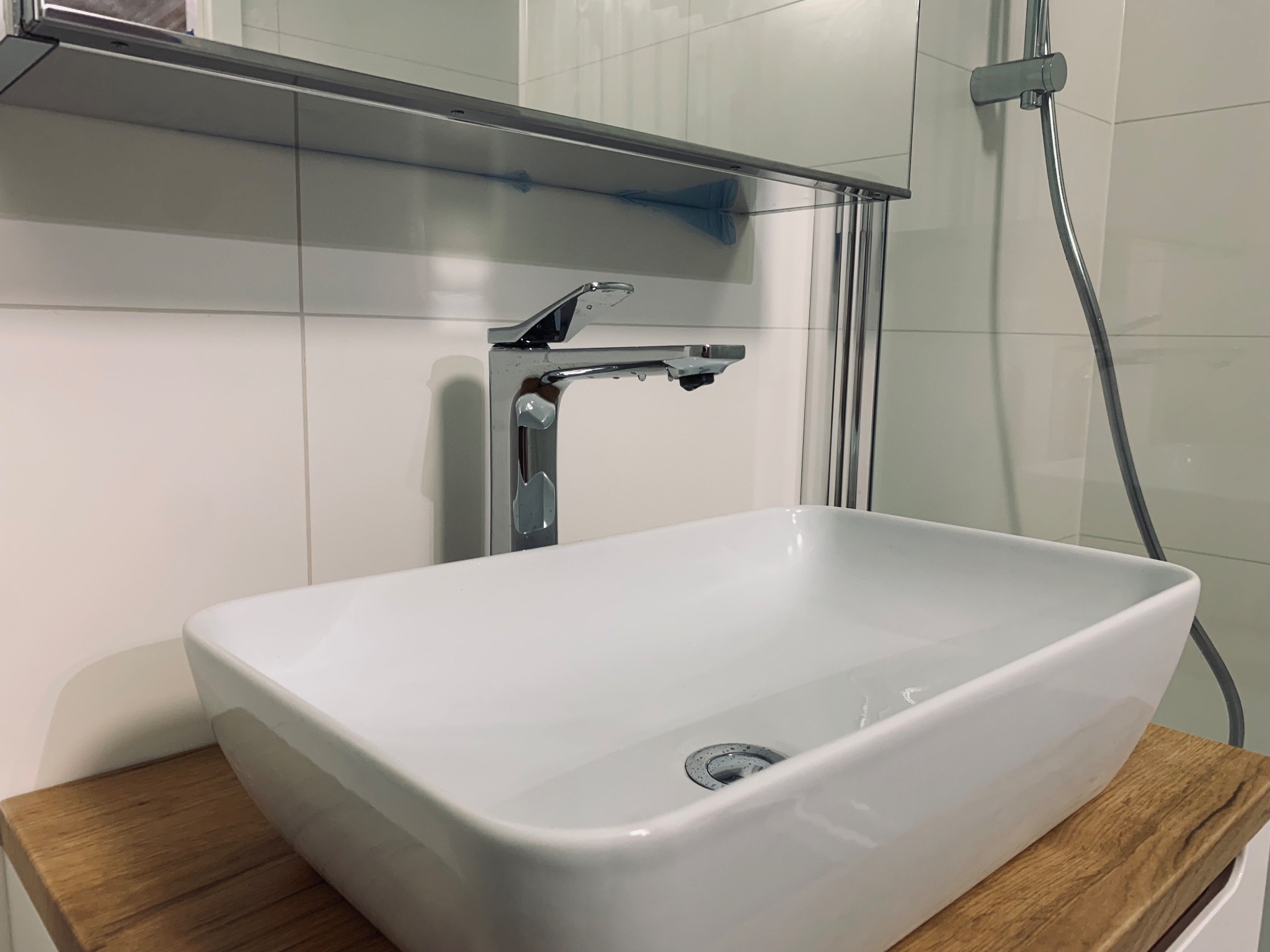 Nowoczesna, prostokątna umywalka nablatowa z chromowaną baterią i odpływem, zamontowana na blacie z drewnopodobnego materiału. W tle widoczne białe płytki i chromowana słuchawka prysznicowa.