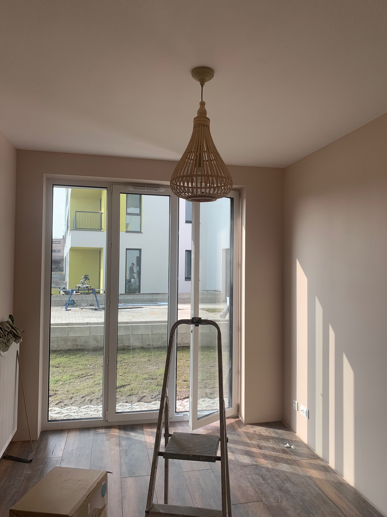 Pokój po malowaniu ścian na delikatny odcień beżu, z widoczną drewnianą lampą wiszącą, drabiną i częściowo ułożonymi panelami podłogowymi w odcieniach brązu, okno z widokiem na budynek w budowie.