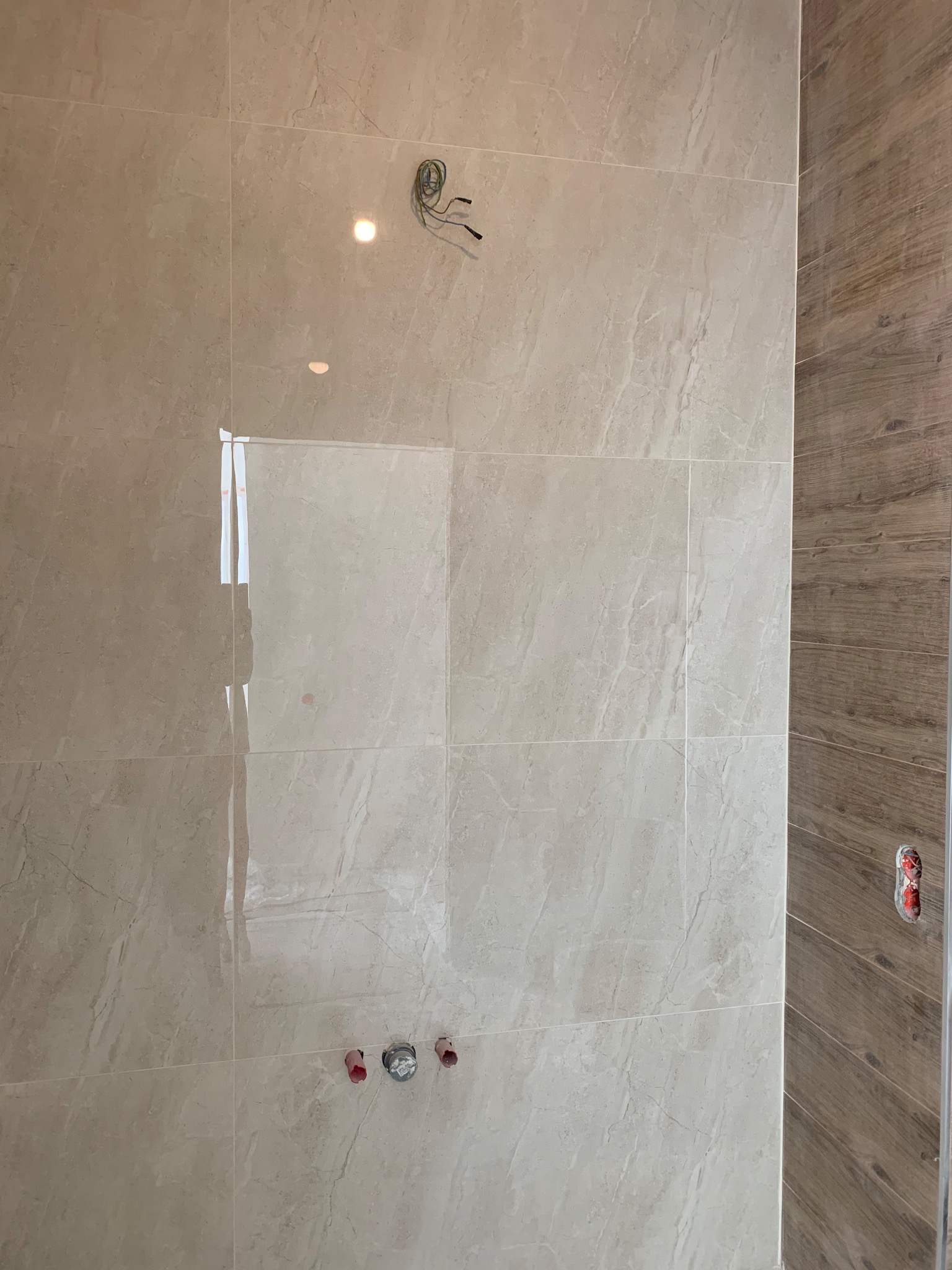 Ściana łazienki wyłożona beżowymi płytkami imitującymi marmur, z widocznymi wystającymi przewodami elektrycznymi i rurami hydraulicznymi przed montażem armatury, obok fragment ściany wyłożony...