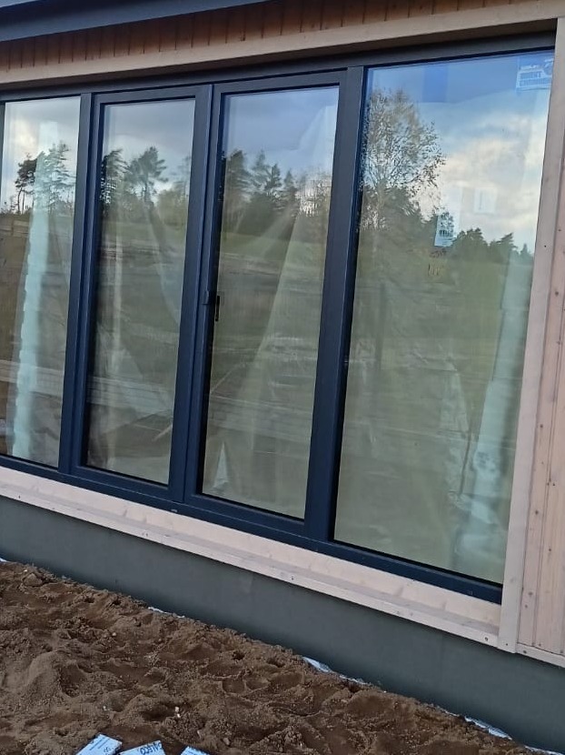 Nowoczesne, grafitowe okna panoramiczne w trakcie montażu w elewacji domu z jasnego drewna, widoczne zabezpieczenia folią i świeżo usypana ziemia.