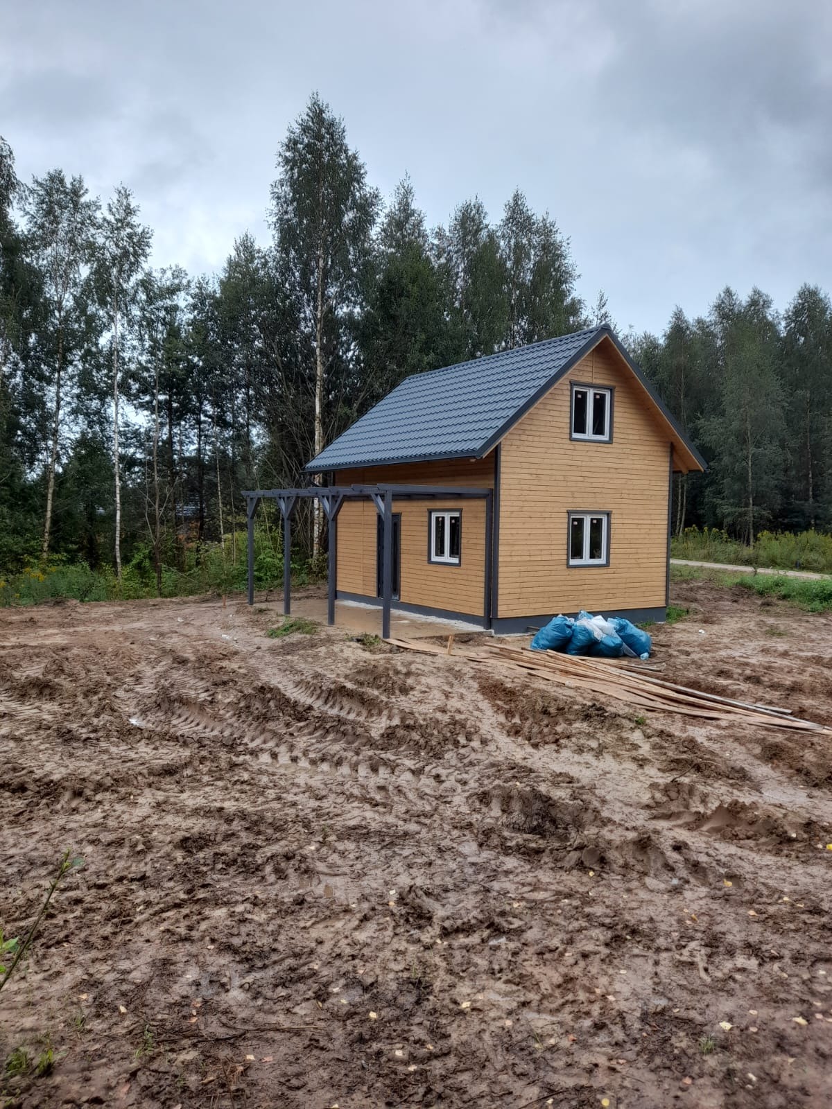 Surowy stan placu budowy z domem szkieletowym z ciemnoszarą dachówką, drewnianą elewacją i zadaszeniem tarasu, widoczne ślady opon w błocie i worki z odpadami.