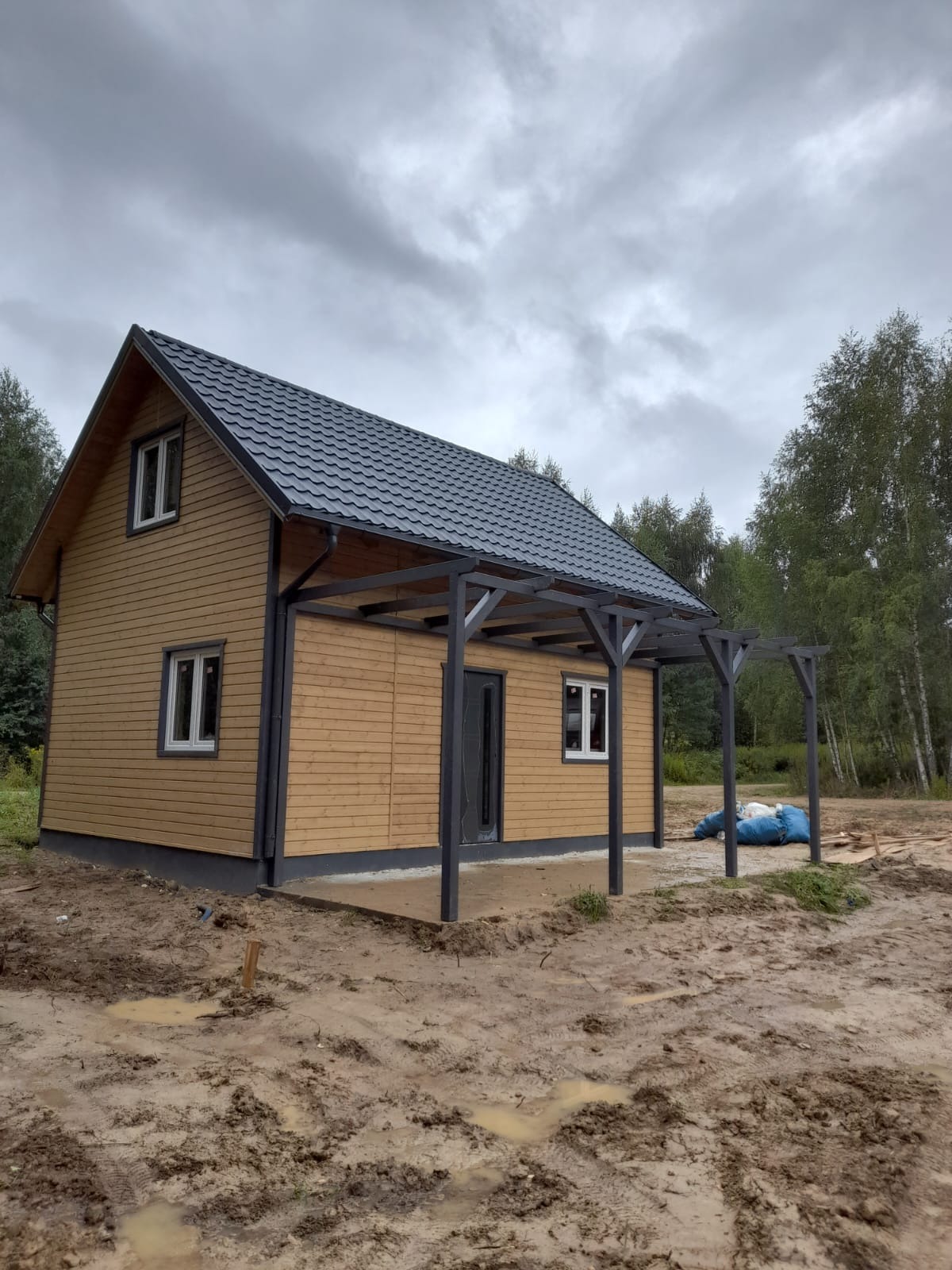 Nowoczesny, parterowy budynek z drewnianą elewacją i ciemnoszarą dachówką, z zadaszeniem tarasowym, na działce z błotnistym podłożem i widocznymi śladami opon.