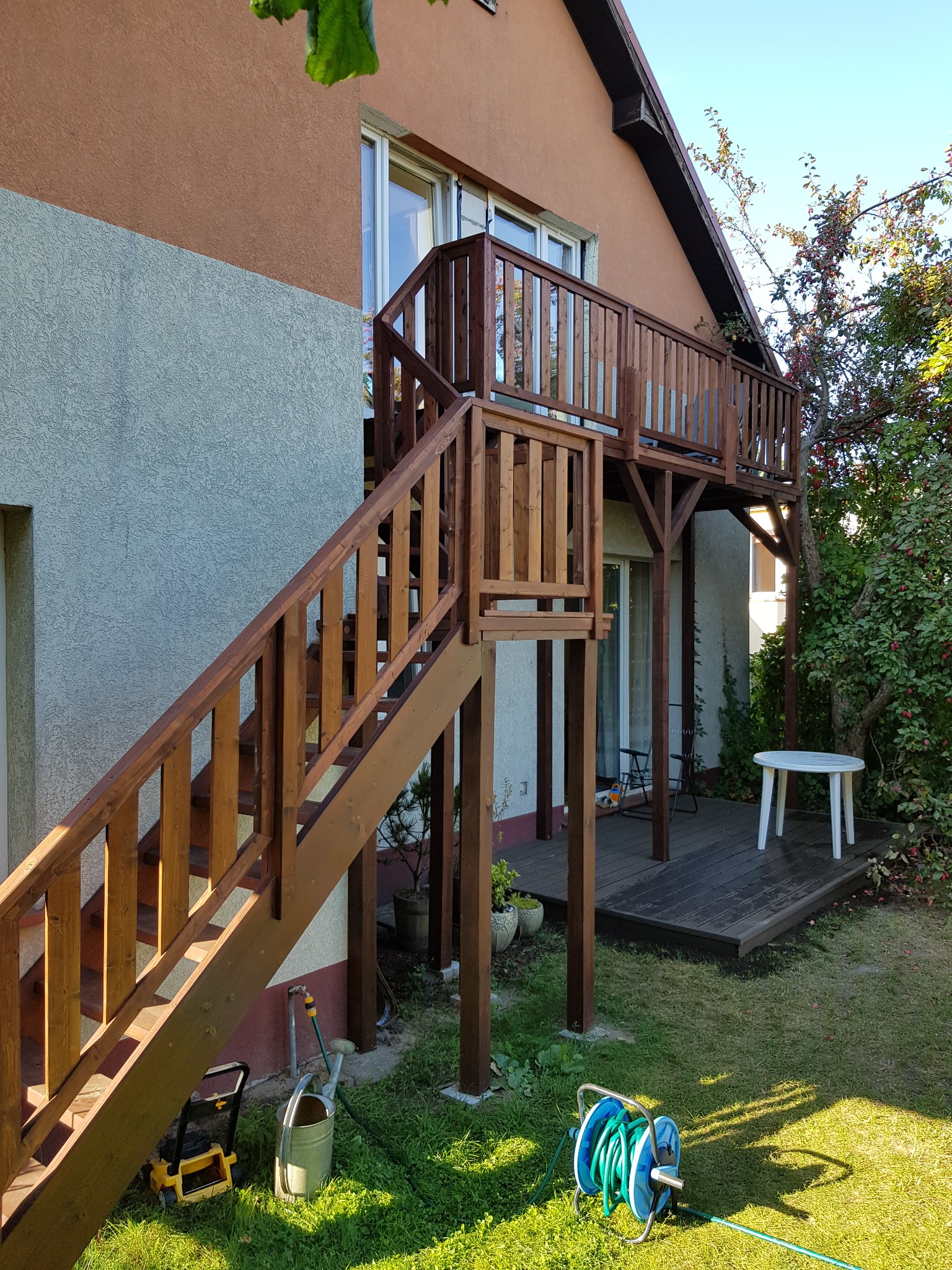 Brązowy drewniany ganek z schodami przylegający do domu z dwukolorową elewacją, na trawniku narzędzia ogrodnicze i zwinięty wąż.