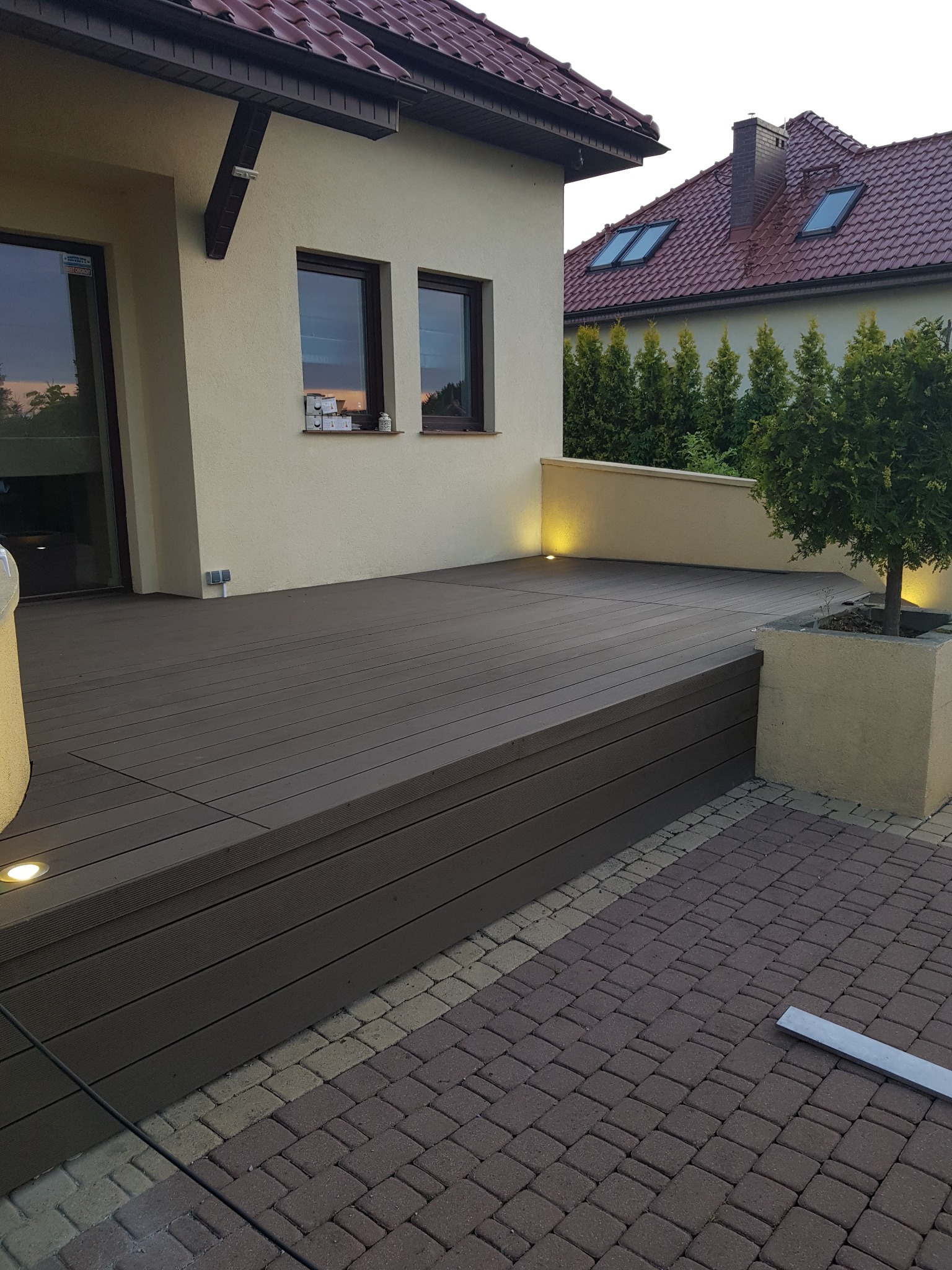 Zewnętrzny taras z desek kompozytowych, z niskim oświetleniem punktowym, przylegający do elewacji budynku z widocznym ogrodem w tle.