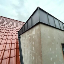 DEKARMAX Jakub Olbryś - Widok z dołu na fragment dachu z czerwoną dachówką ceramiczną i fragment budynku z szarą, metalową obróbką blacharską na dachu, ściana budynku w trakcie ocieplania, widoczne siatki i klej.