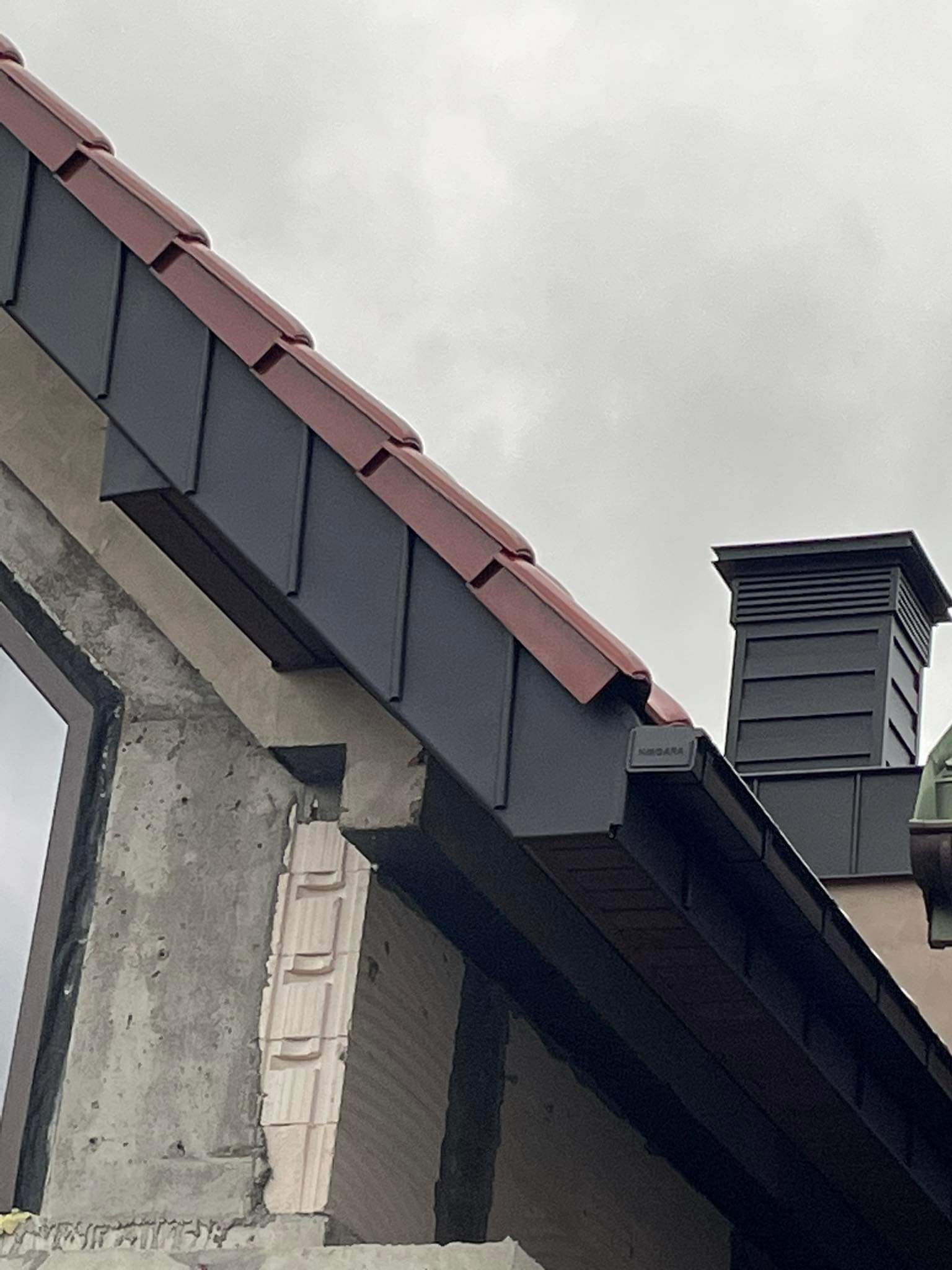 Fragment dachu z ciemnoszarą blachą, czerwona dachówka przy krawędzi, widoczny komin i fragment ściany budynku w trakcie budowy, pochmurne niebo w tle.