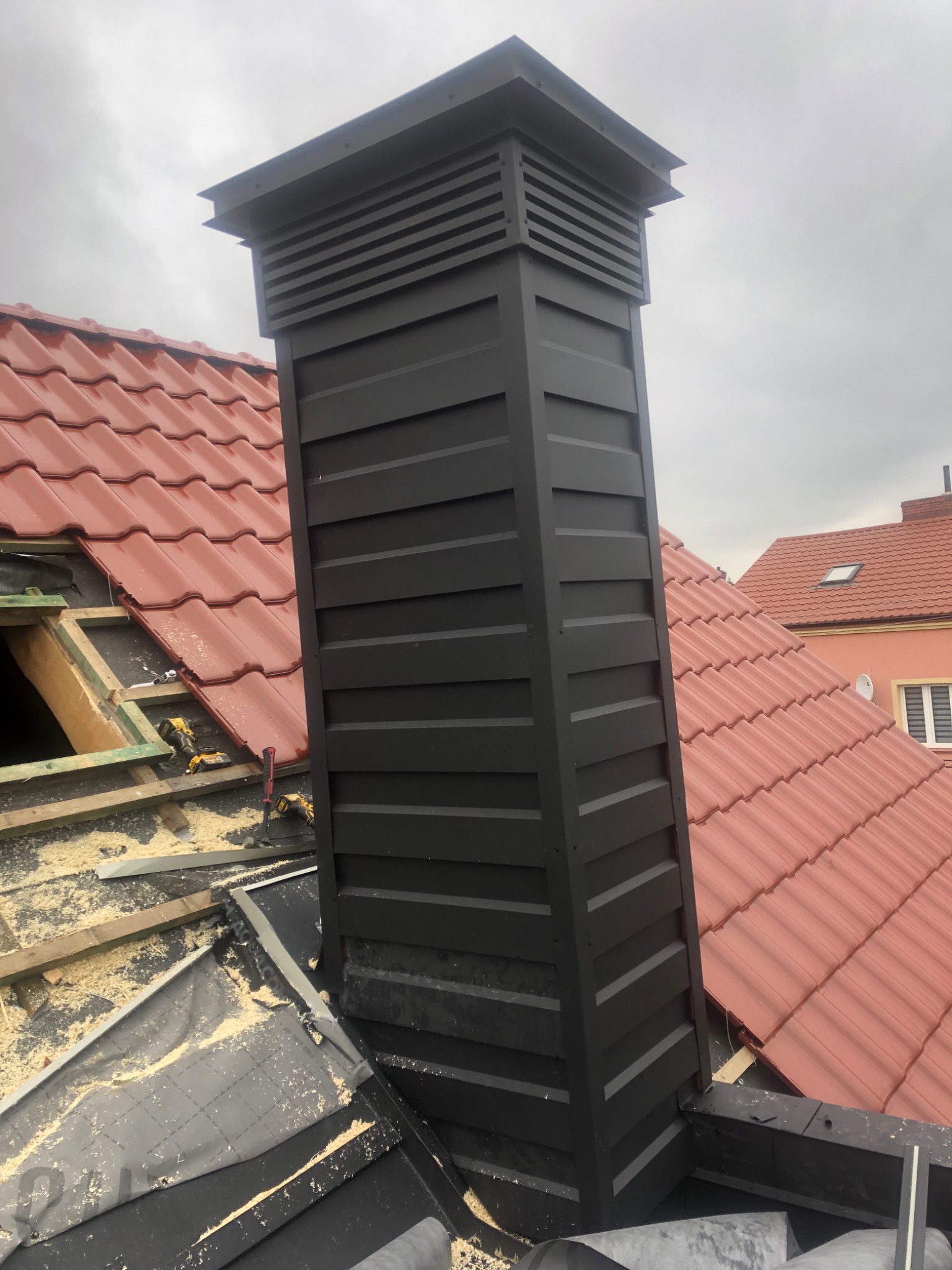 Czarny komin wentylacyjny na dachu z czerwonej dachówki, widoczne narzędzia i elementy konstrukcyjne dachu w trakcie montażu.