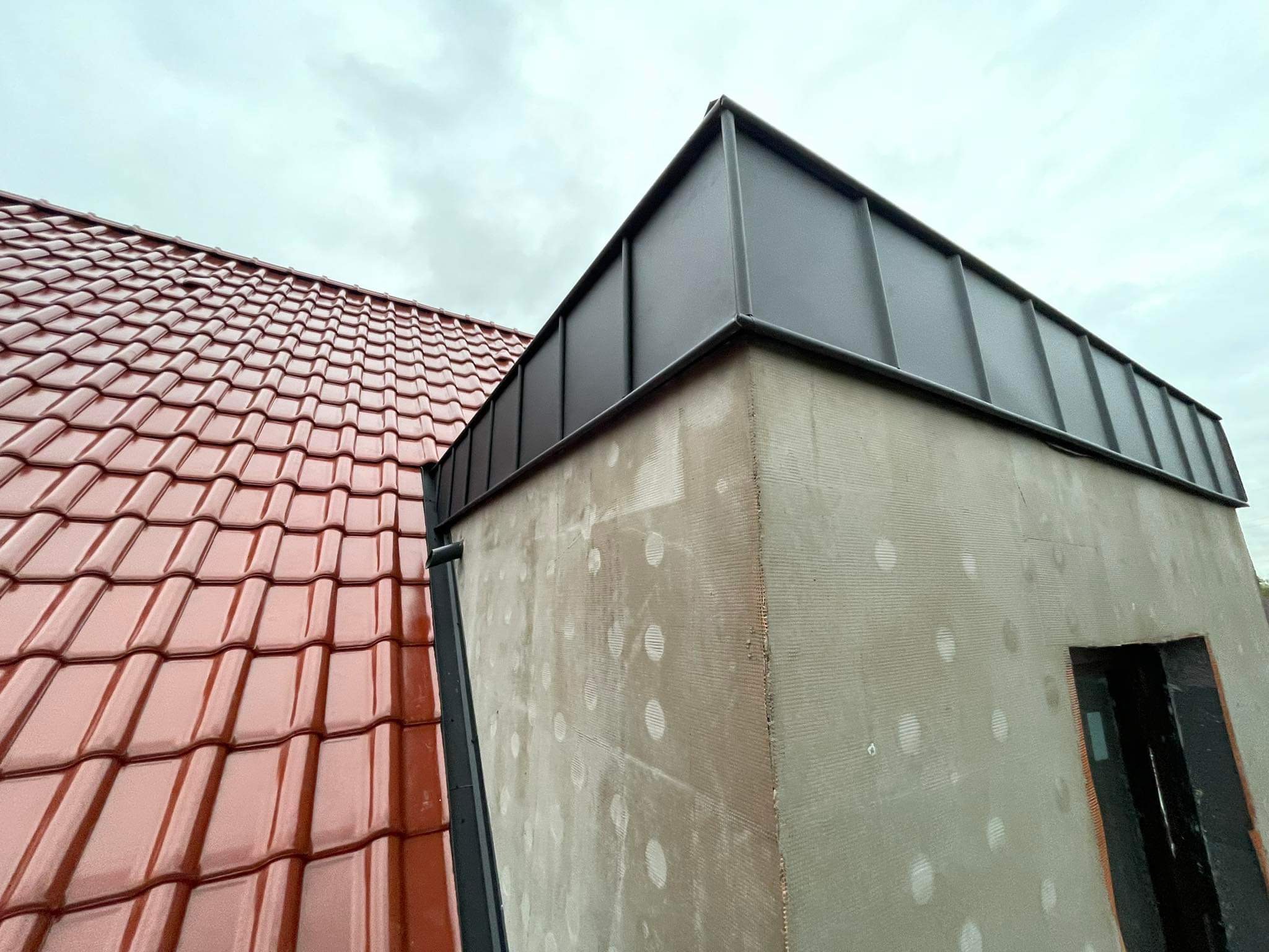 Widok z dołu na fragment dachu z czerwoną dachówką ceramiczną i fragment budynku z szarą, metalową obróbką blacharską na dachu, ściana budynku w trakcie ocieplania, widoczne siatki i klej.