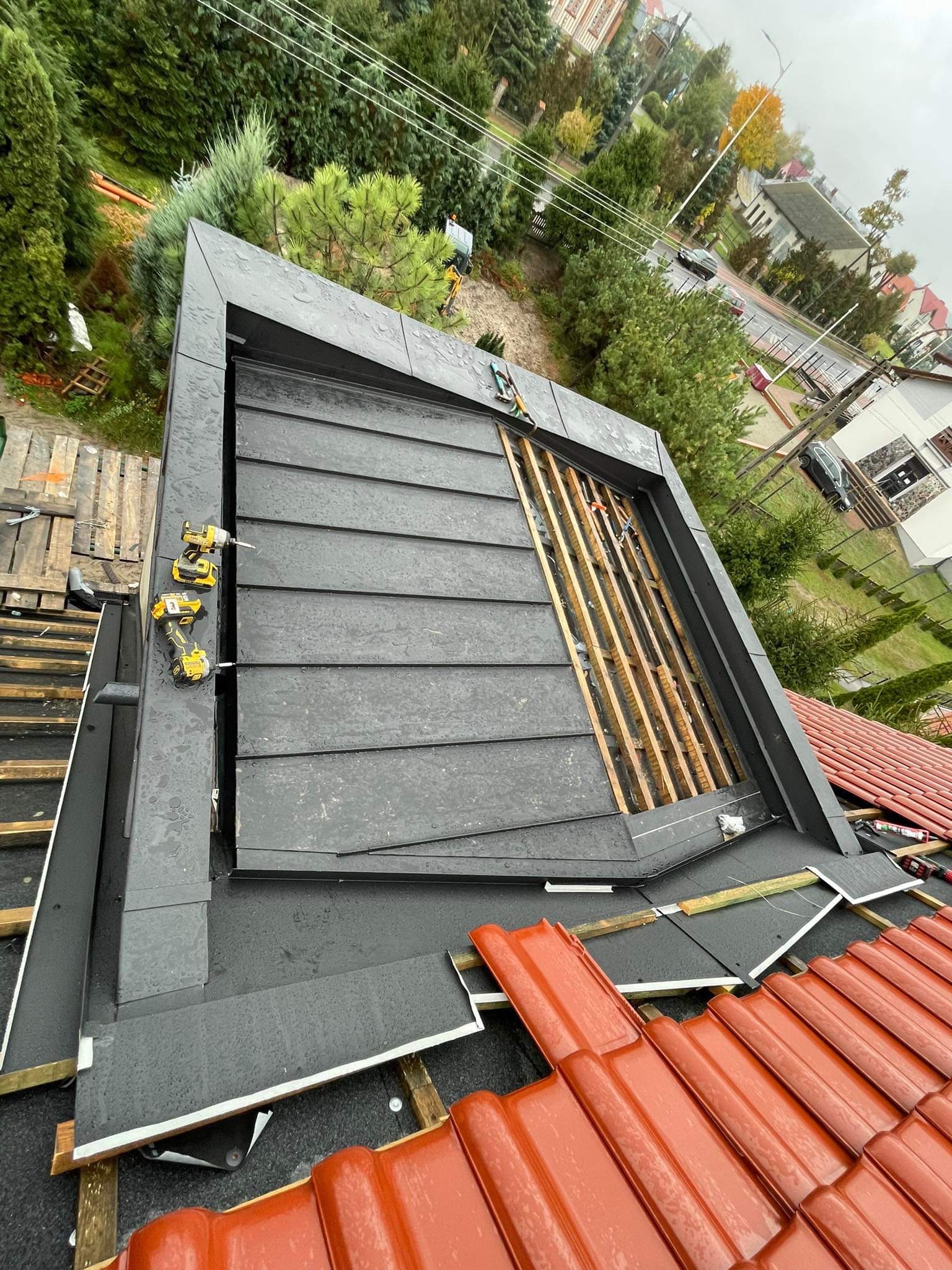 Montaż obróbek blacharskich na dachu z widoczną więźbą i narzędziami, ujęcie z góry w pochmurny dzień.