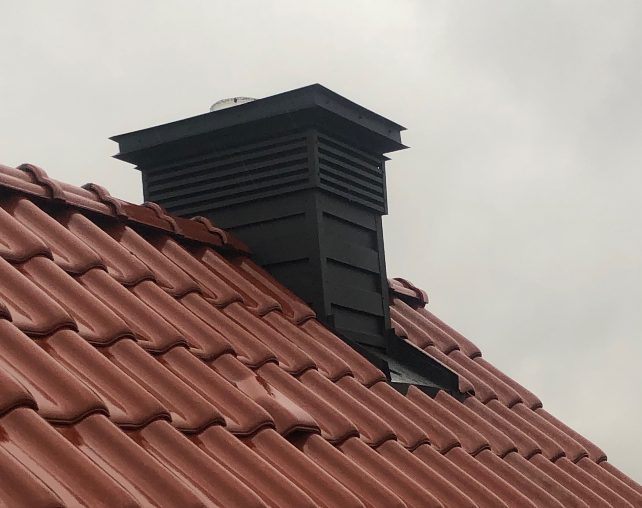 Czerwona dachówka ceramiczna z czarnym kominem wentylacyjnym, widok z dołu na tle pochmurnego nieba.