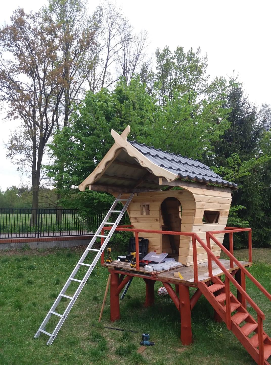 Konstrukcja drewnianego domku dla dzieci z czarną dachówką, na podwyższeniu z czerwonymi barierkami i schodkami, w trakcie budowy, z drabiną opartą o dach i narzędziami na podeście.