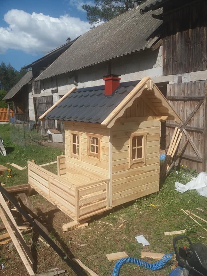 Niedokończony drewniany domek dla dzieci z czarnym dachem i czerwonym kominem, w tle budynki gospodarcze z szarym dachem z eternitu, narzędzia i fragmenty drewna wokół domku.