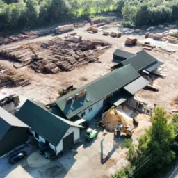 Widok z lotu ptaka na teren tartaku z budynkami, stosem pni drzew, tarcicy i zrębków, maszynami budowlanymi oraz zaparkowanymi samochodami osobowymi.