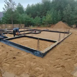 Przygotowanie szalunku fundamentów pod budowę domu jednorodzinnego, widoczni dwaj pracownicy przy pracy, teren budowy z piaskiem i drzewami w tle.