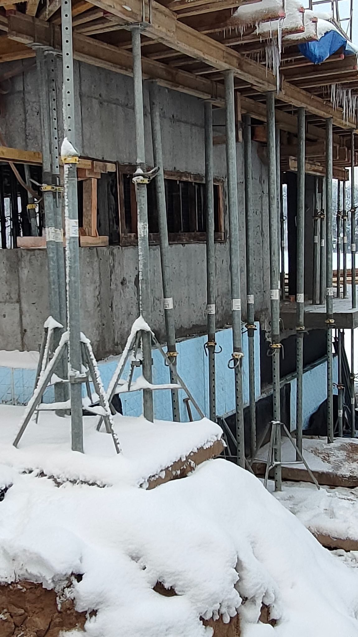 Surowy betonowy budynek w trakcie budowy, wsparty metalowymi stemplami i rusztowaniami, częściowo ocieplony niebieskim styropianem, pokryty śniegiem i zwisającymi soplami lodu.