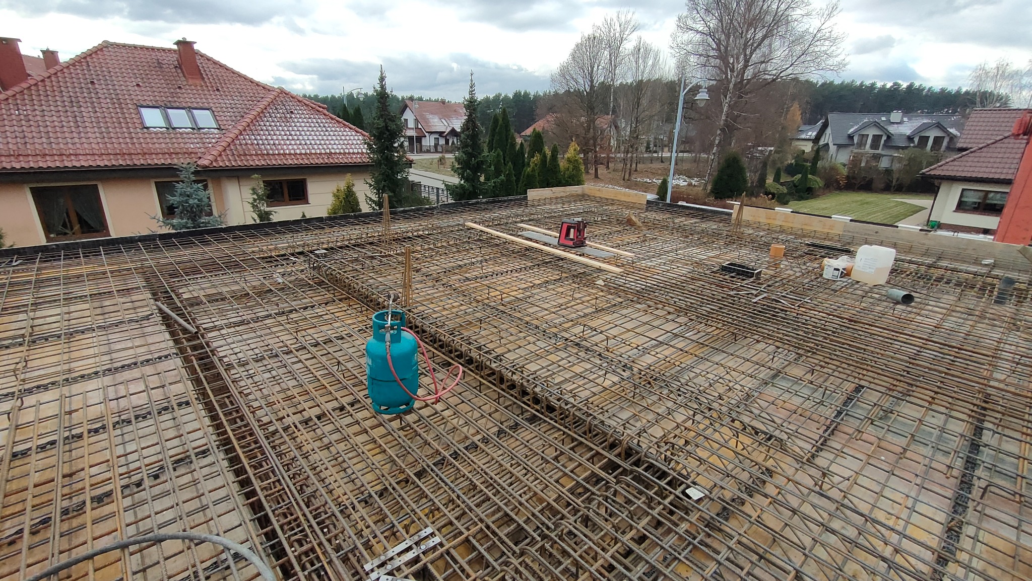 Siatka zbrojeniowa na fundamencie budynku, widoczna butla gazowa i poziomica laserowa, w tle zabudowa jednorodzinna.
