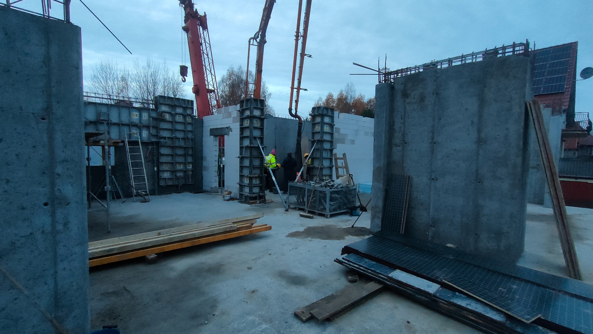 Budowa domu: betonowe ściany szalunkowe w trakcie wylewania, widoczne pompy do betonu, pracownicy w kamizelkach, niebo pochmurne.