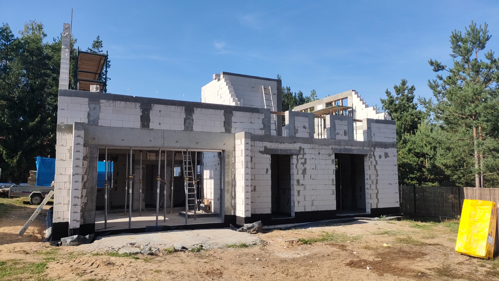 Wznoszenie dwukondygnacyjnego budynku z bloczków, widoczne szalunki nad otworami okiennymi i drzwiowymi, rusztowanie z boku budynku, teren budowy w otoczeniu drzew