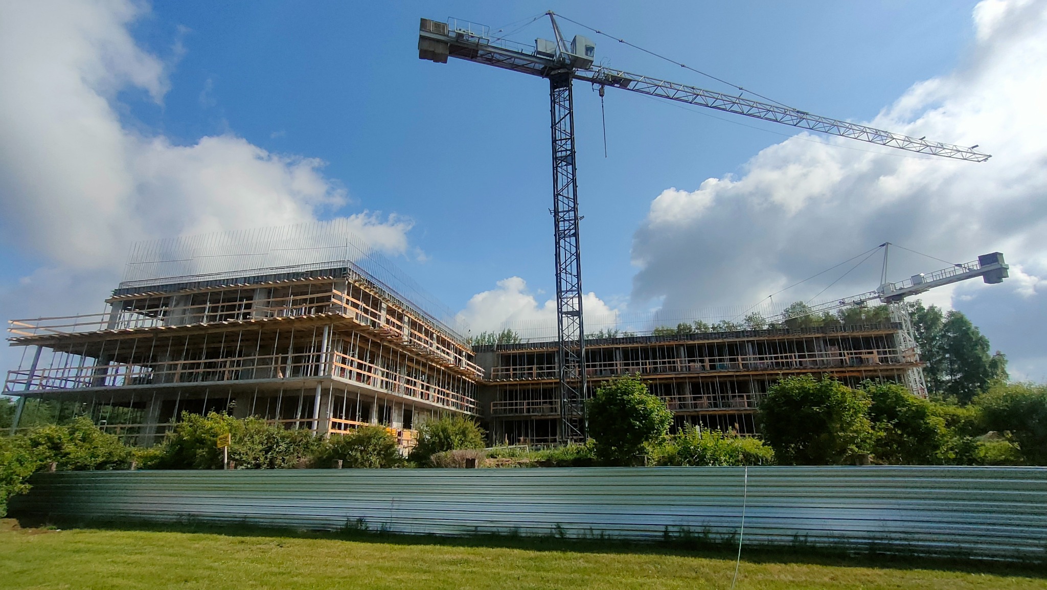 Budowa wielopiętrowego budynku z betonu i drewna, widoczne rusztowania i dwa żurawie wieżowe na tle błękitnego nieba z chmurami, ogrodzenie z blachy falistej w pierwszym planie.