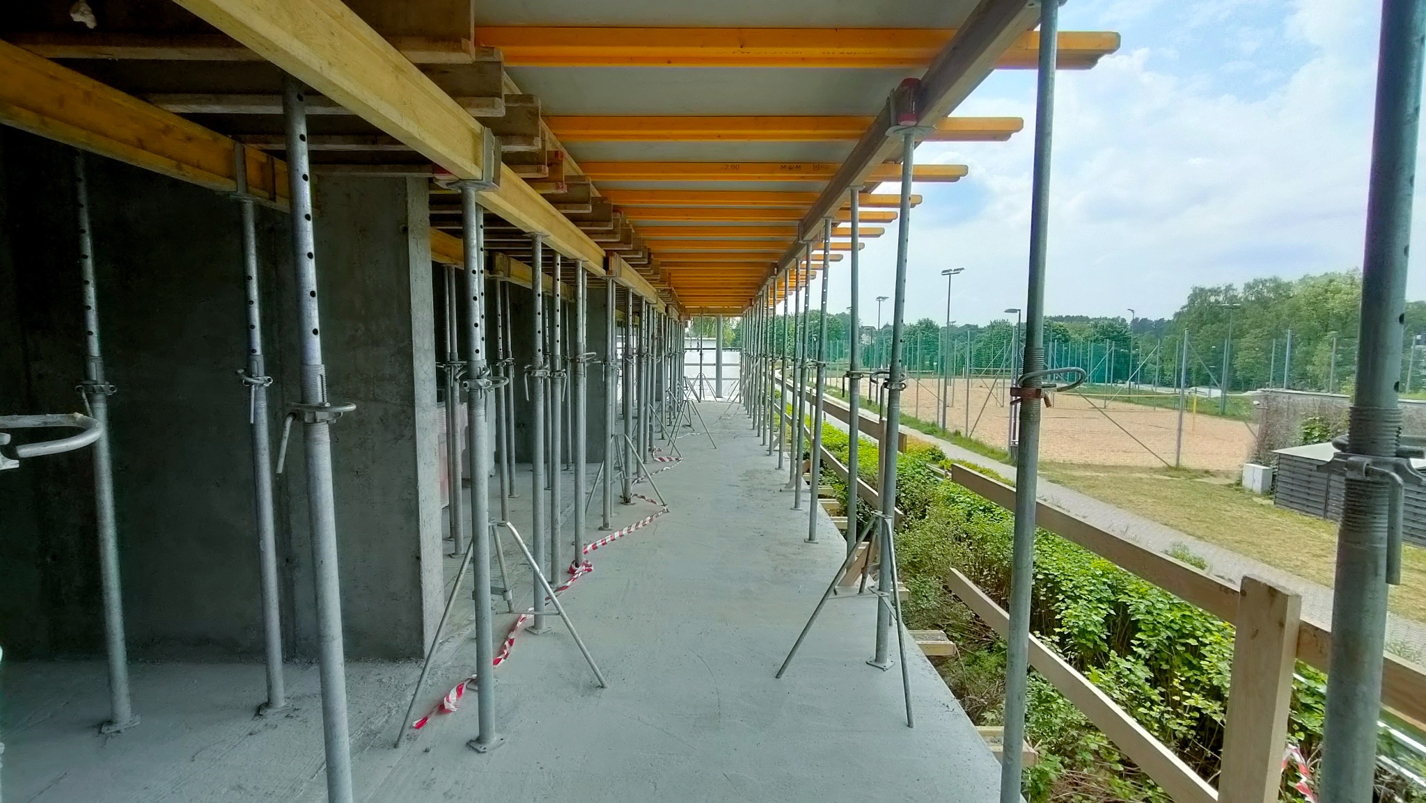 Szare betonowe ściany i strop w trakcie budowy, podparte metalowymi stemplami budowlanymi, widok na zewnątrz na boisko sportowe i zieleń.