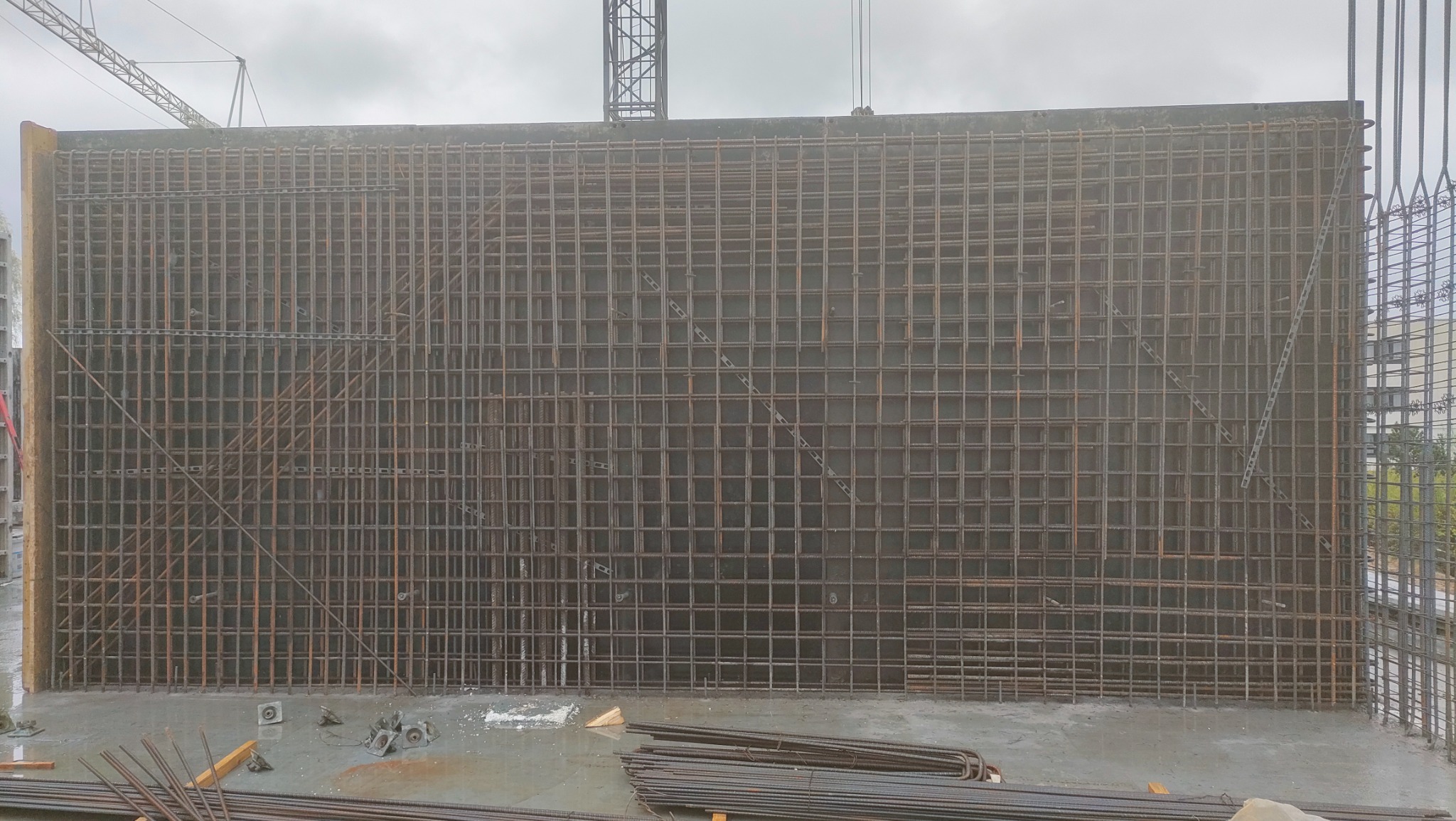 Zbrojenie pionowej ściany szalunkowej z widocznymi elementami montażowymi i prętami na mokrym betonie, w tle dźwig budowlany.