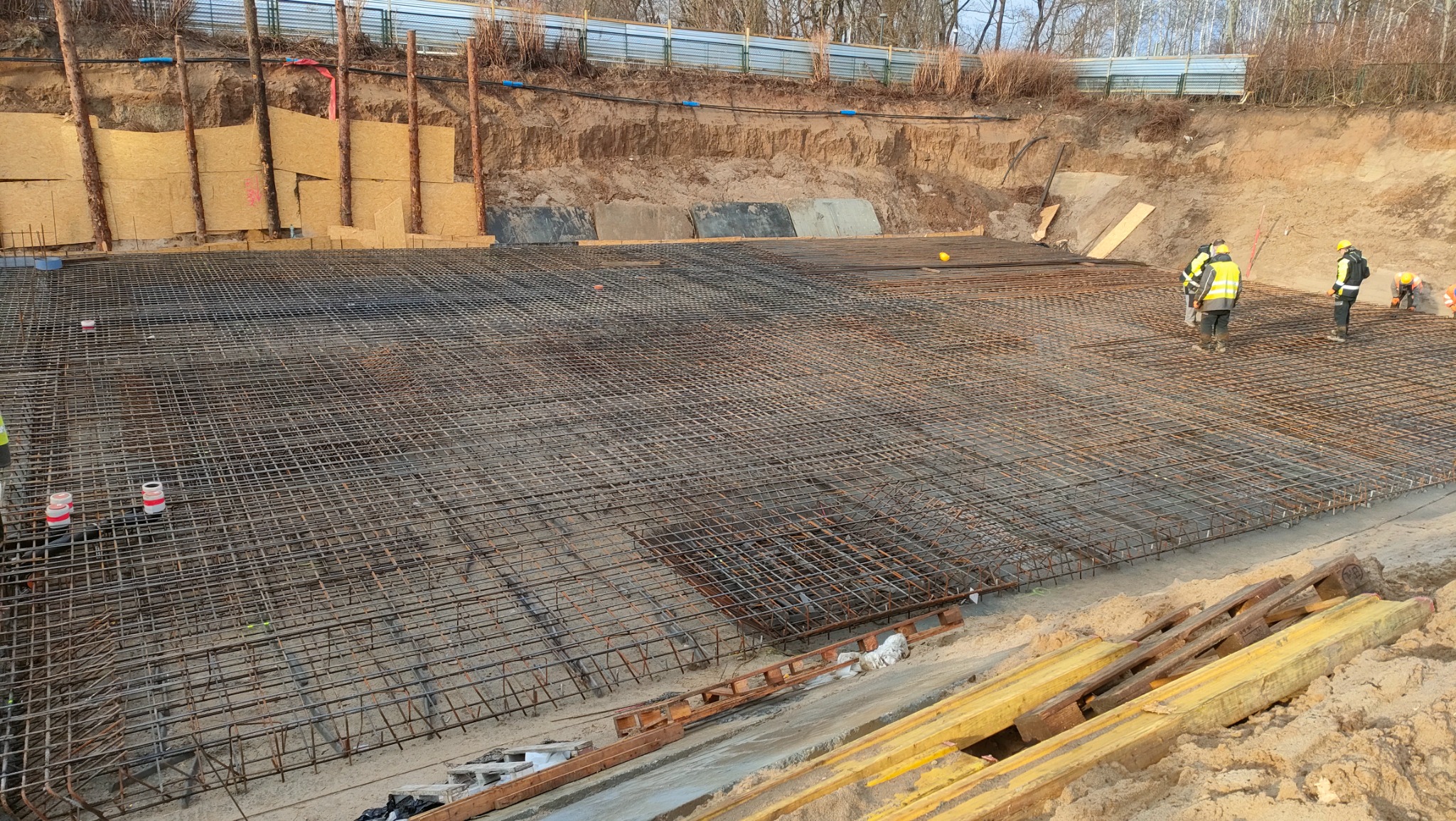 Zbrojenie płyty fundamentowej z widocznymi pracownikami w odblaskowych kamizelkach, zabezpieczone wykopu deskowaniem i ziemią.