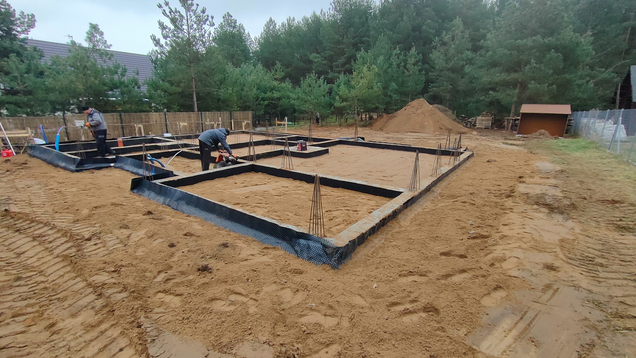 Przygotowanie szalunku fundamentów pod budowę domu jednorodzinnego, widoczni dwaj pracownicy przy pracy, teren budowy z piaskiem i drzewami w tle.