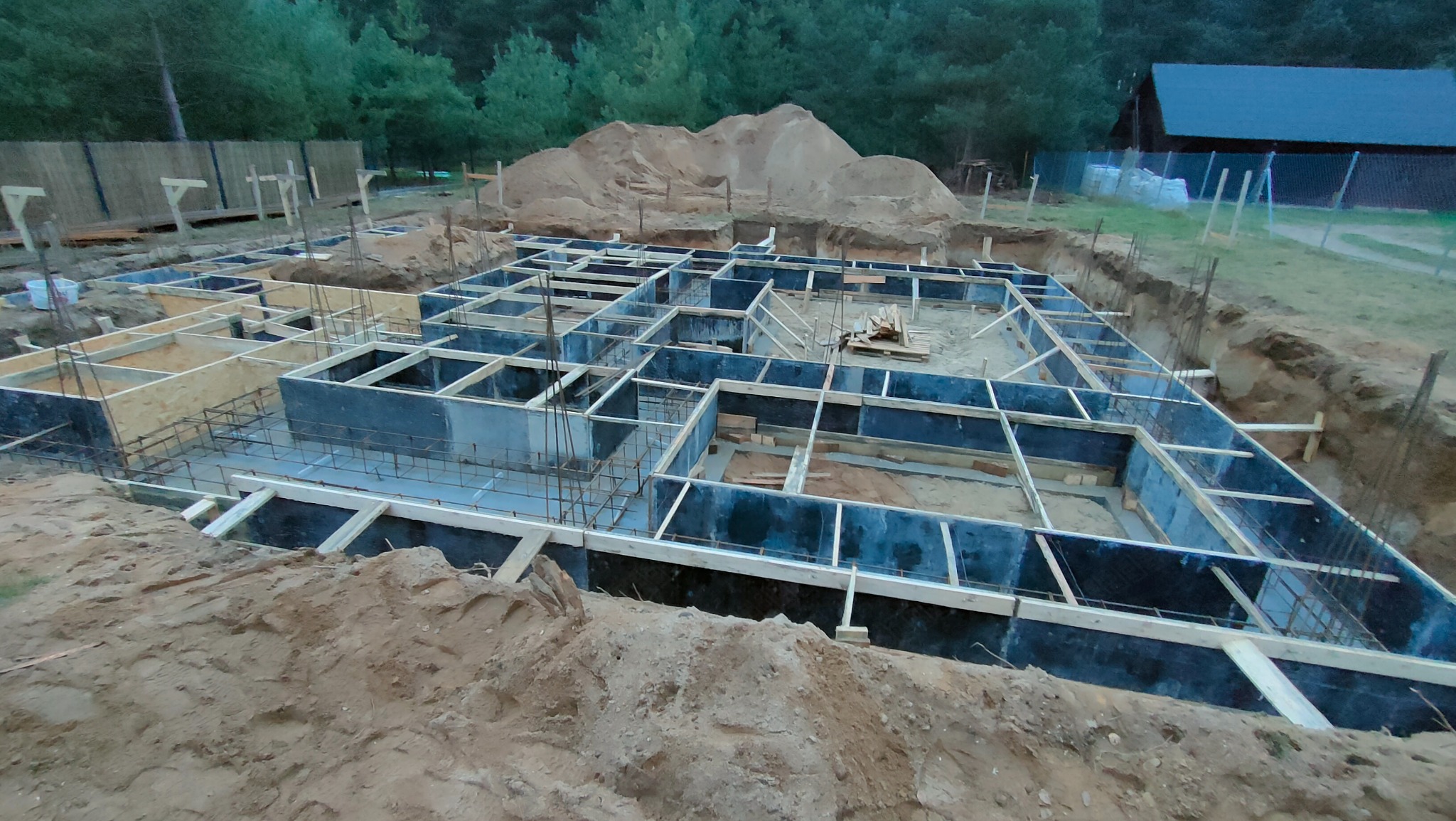Zalany beton w szalunkach tworzących fundament domu jednorodzinnego, wystające pręty zbrojeniowe, w tle pryzma piasku i zalesiony teren.