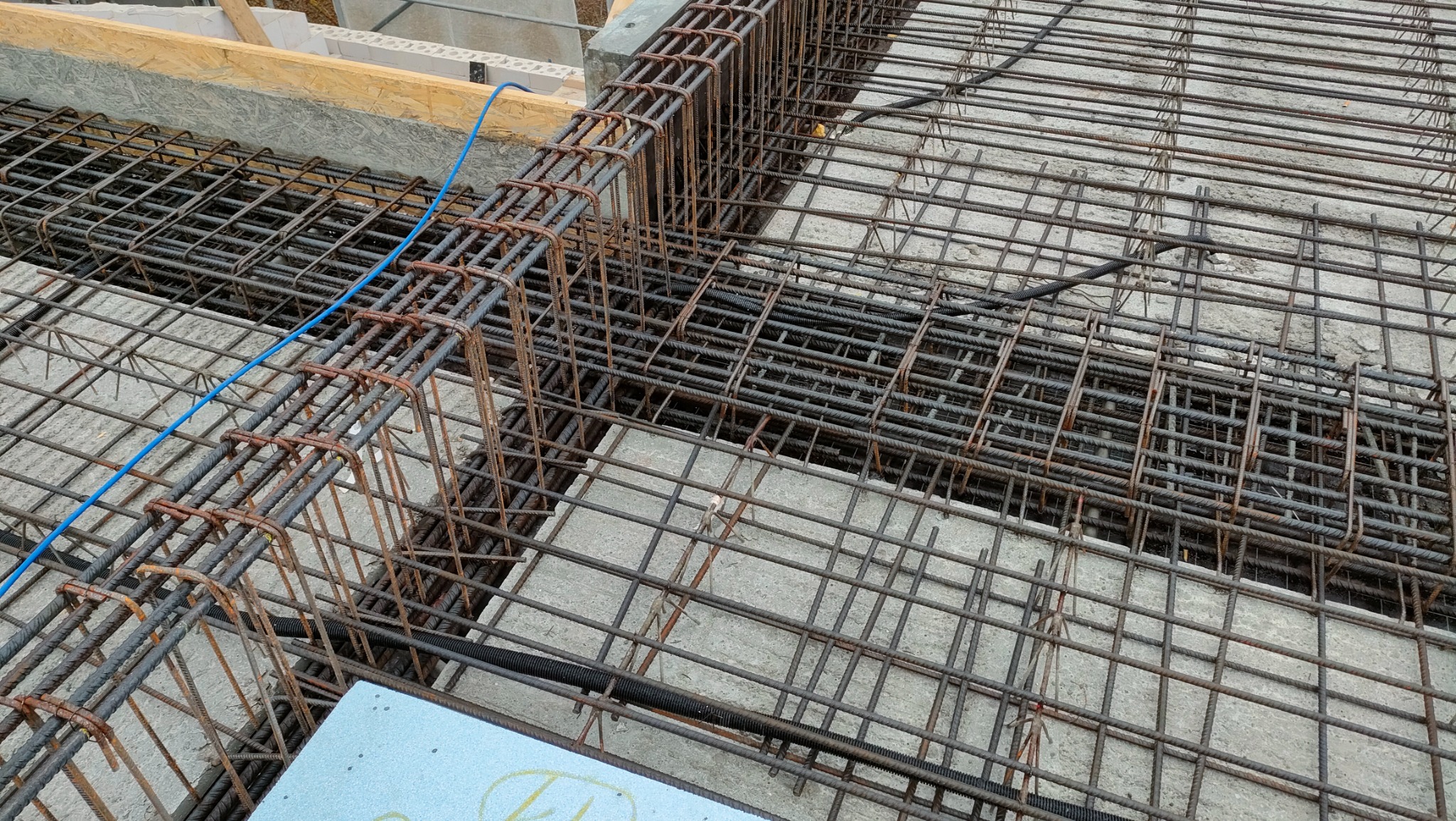 Zbrojenie fundamentów z widocznymi prętami pionowymi i poziomymi, częściowo zalane betonem, z niebieskim przewodem i styropianem w rogu kadru.