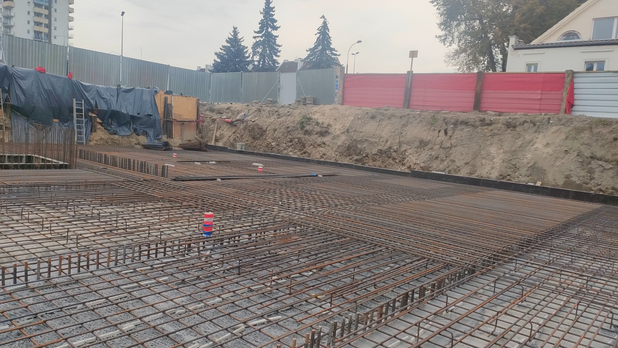 Zbrojenie płyty fundamentowej na placu budowy, widoczne stalowe pręty tworzące siatkę, w tle ogrodzenie placu budowy i budynki.