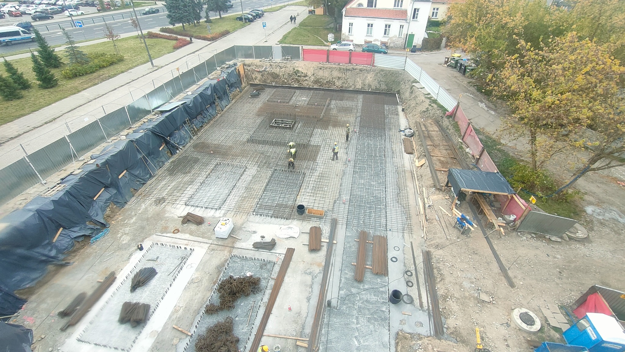 Widok z góry na plac budowy z wykopem pod fundamenty, gdzie robotnicy w kaskach układają zbrojenie; w tle widoczna ulica i budynki.