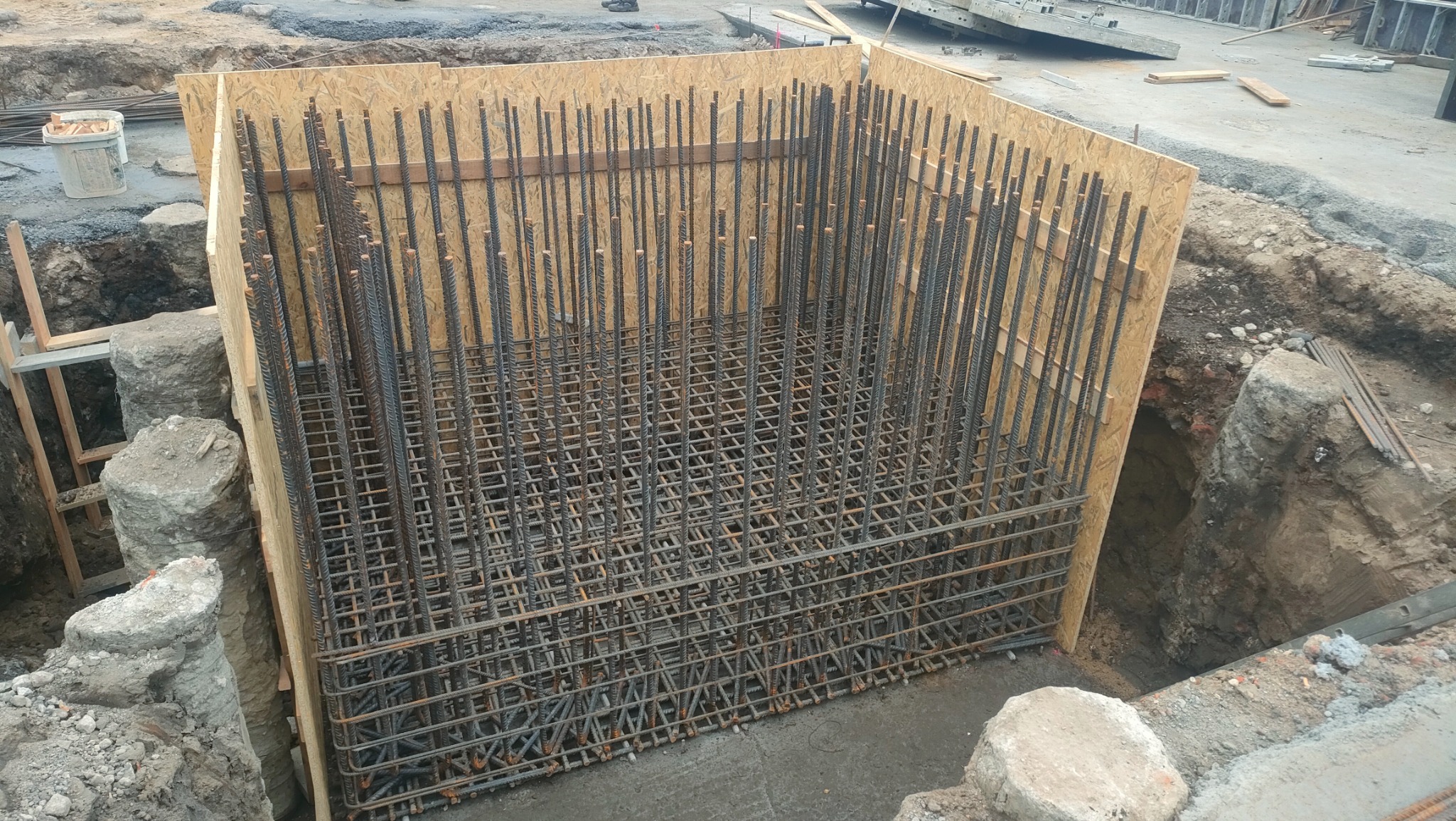 Zbrojenie fundamentu w drewnianym szalunku, widoczne wystające pręty pionowe i poziome kratownice, otoczone wykopem w ziemi.