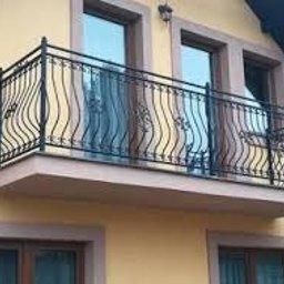 Mmetal - Czarna, metalowa balustrada balkonowa z dekoracyjnymi, falistymi elementami, zamontowana na balkonie budynku o żółtej elewacji. Widoczne okna z brązowymi ramami.