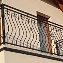 Mmetal - Czarna, metalowa balustrada balkonowa z dekoracyjnymi, falistymi prętami, zamontowana na jasnej elewacji budynku.