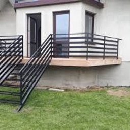 Mmetal - Zewnętrzne schody metalowe z czarną, poziomą balustradą prowadzące do jasnego wejścia domu, widoczne trawnik i fragment elewacji.