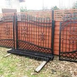 Mmetal - Elementy kutego ogrodzenia w kolorze czarnym, oparte o drewniane panele ogrodzeniowe, z widocznym fragmentem trawnika i domu w tle.