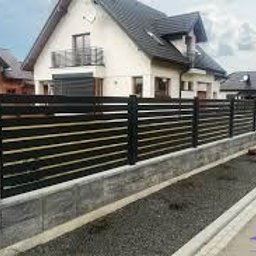 Mmetal - Nowoczesne ogrodzenie posesji z poziomych, ciemnych desek na betonowej podmurówce i słupkach z szarego betonu architektonicznego, dom w tle.