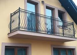 Czarna, metalowa balustrada balkonowa z dekoracyjnymi, falistymi elementami, zamontowana na balkonie budynku o żółtej elewacji. Widoczne okna z brązowymi ramami.